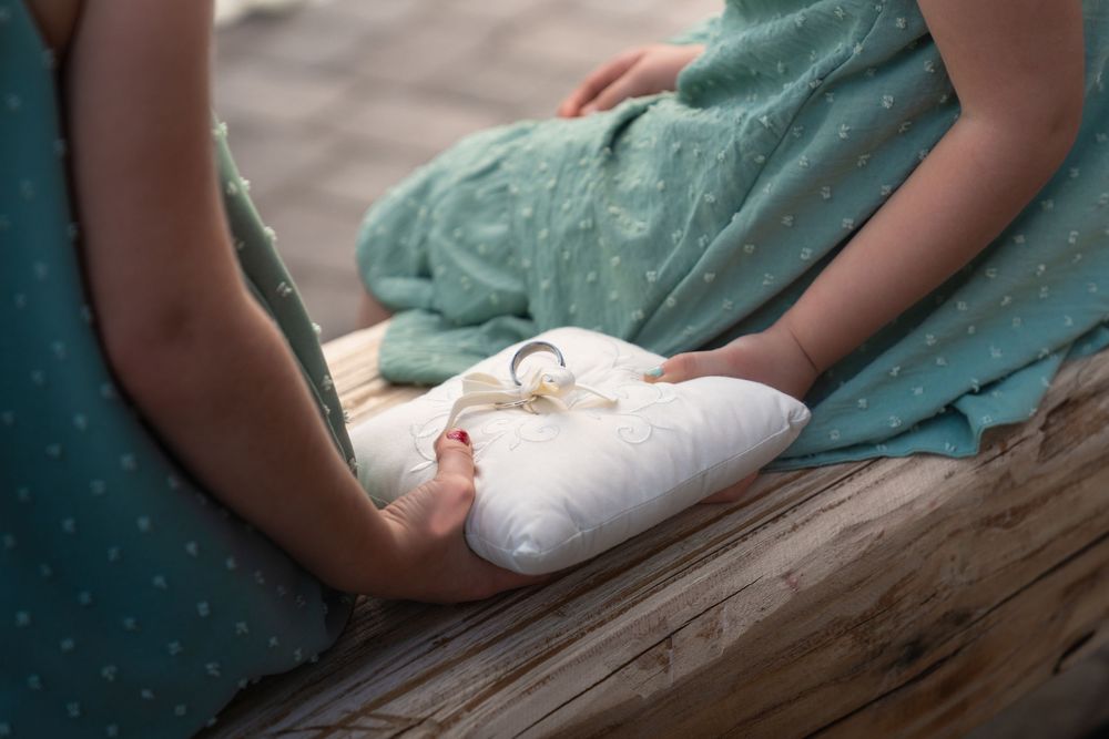 Wedding rings rest on a white pillow held by hands in a teal dress.