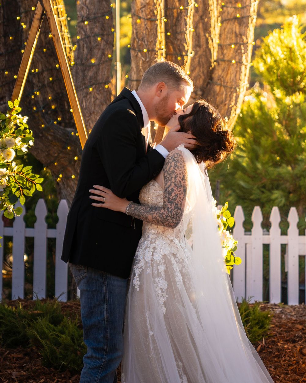 Newlyweds embrace near a white picket fence during golden hour with sunlit trees in background.