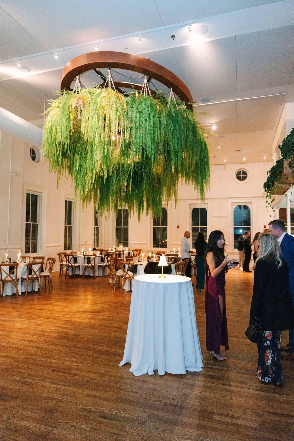 Elegant event space with hanging green willow-style decorations and white round tables on hardwood floors.