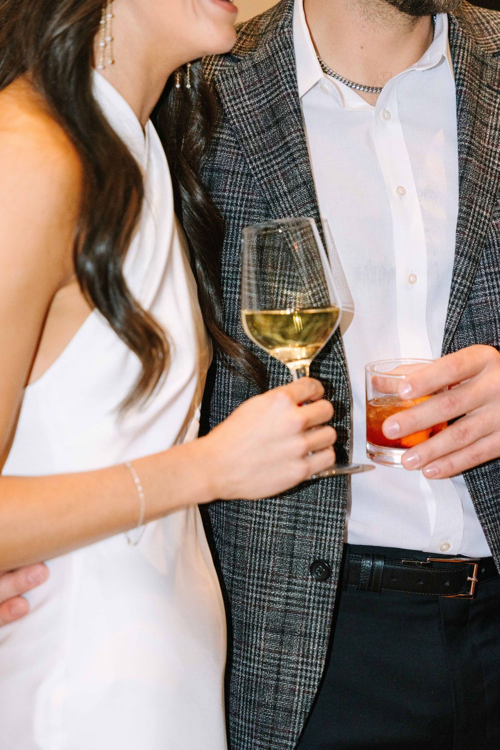 A couple in formal attire holds cocktails, with the woman in white and man wearing a plaid blazer.