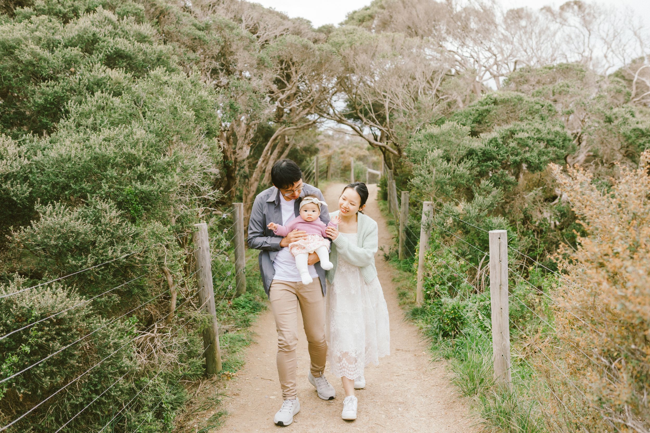 A family walks together on a scenic dirt path surrounded by lush green trees and foliage on a sunny day.
