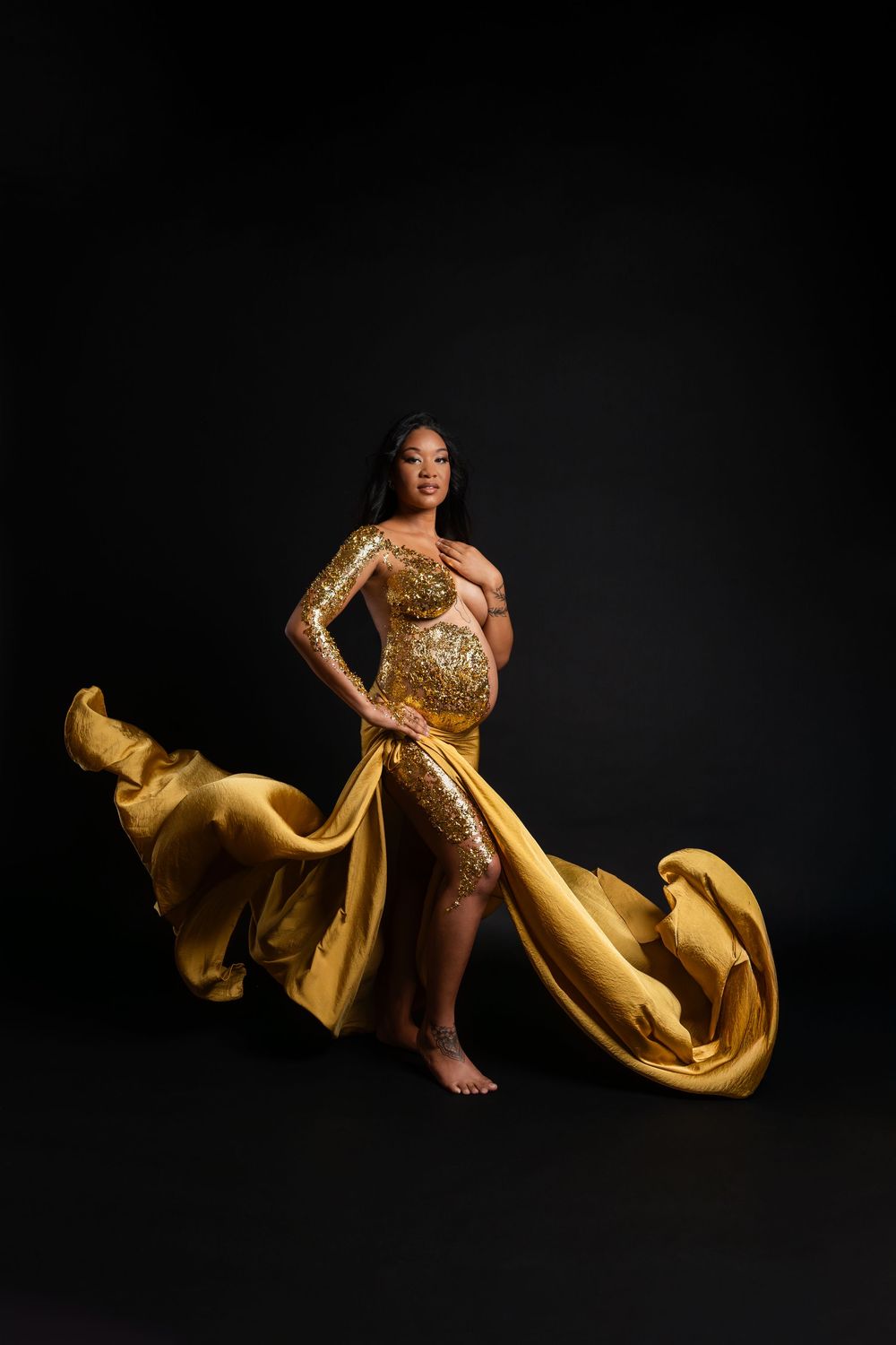 Elegant maternity portrait featuring a flowing gold sequined gown against a dark background with dramatic fabric movement.