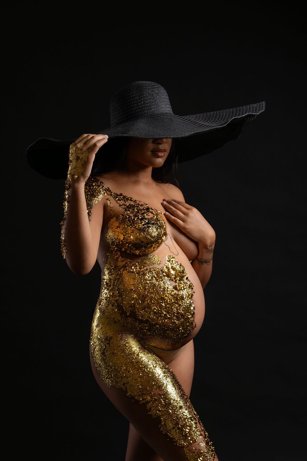 Dramatic maternity portrait with gold body paint and black wide-brimmed hat in dark studio setting.