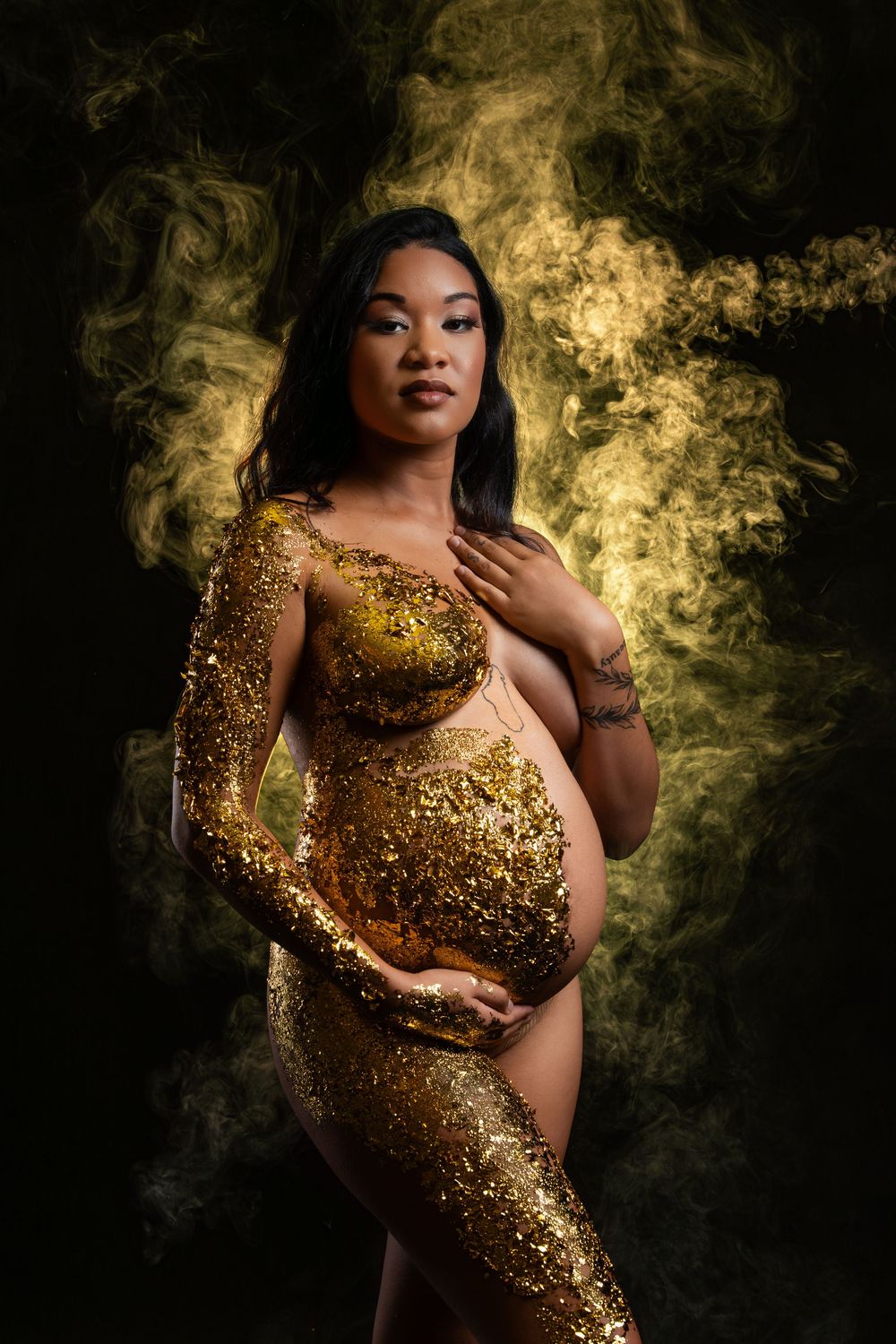 Dramatic maternity photo with gold glitter bodysuit surrounded by ethereal golden smoke effects.