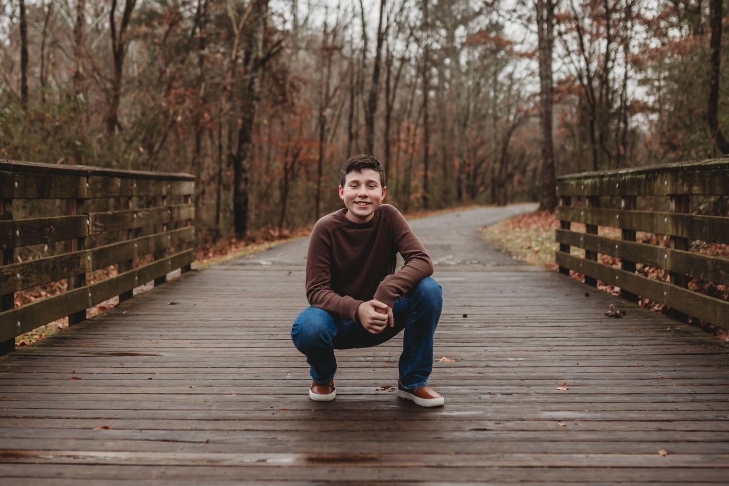 A person sits and poses on a wooden bridge surrounded by autumn trees on a cloudy day.