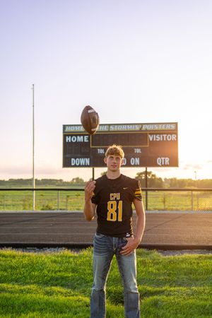 Football player in brown jersey number 81 poses in front of scoreboard during golden hour sunset on athletic field.