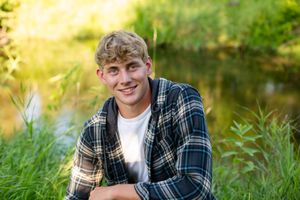 High school senior poses outdoors in a plaid flannel shirt against a natural green background in golden sunlight.