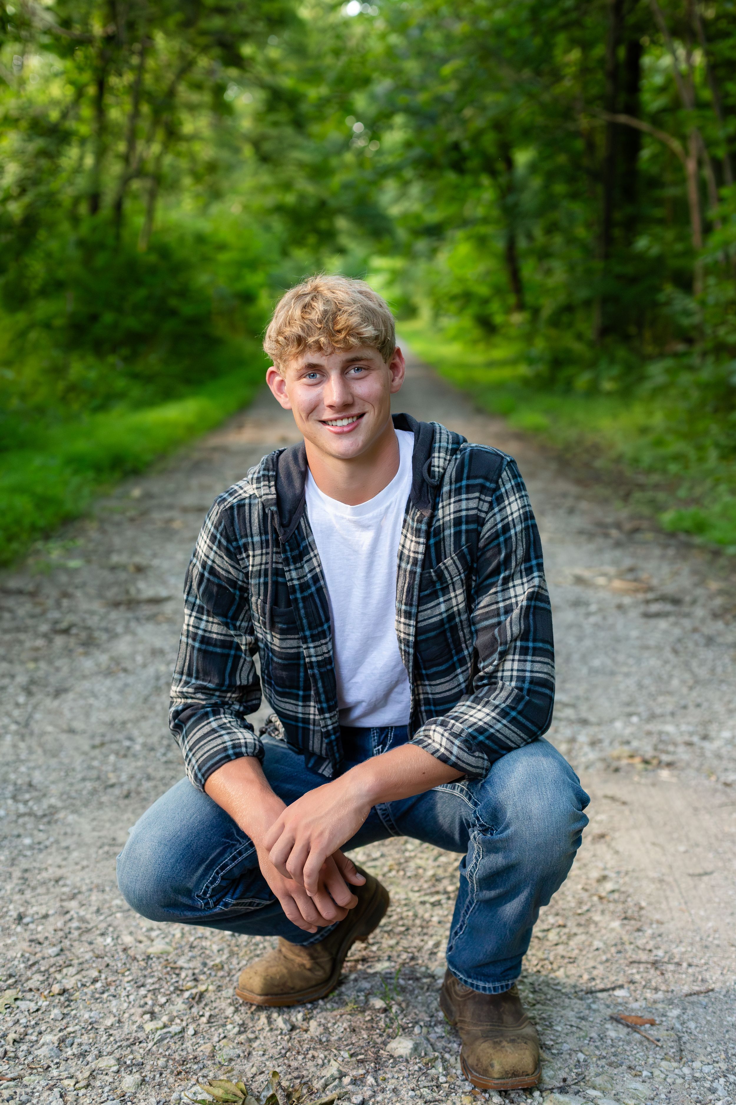 A person wearing a plaid shirt and jeans crouches on a gravel path surrounded by lush green forest.