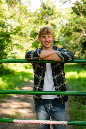 A person in a plaid flannel shirt and jeans leans against green railings in a sunny outdoor park setting.