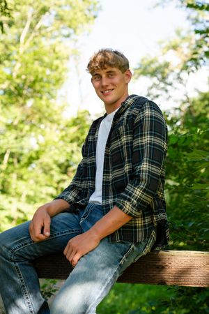 A person in a plaid flannel shirt and jeans sits casually on a wooden fence surrounded by green foliage.