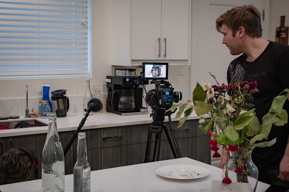 Film set with professional cameras, flowers and water bottles on kitchen counter.