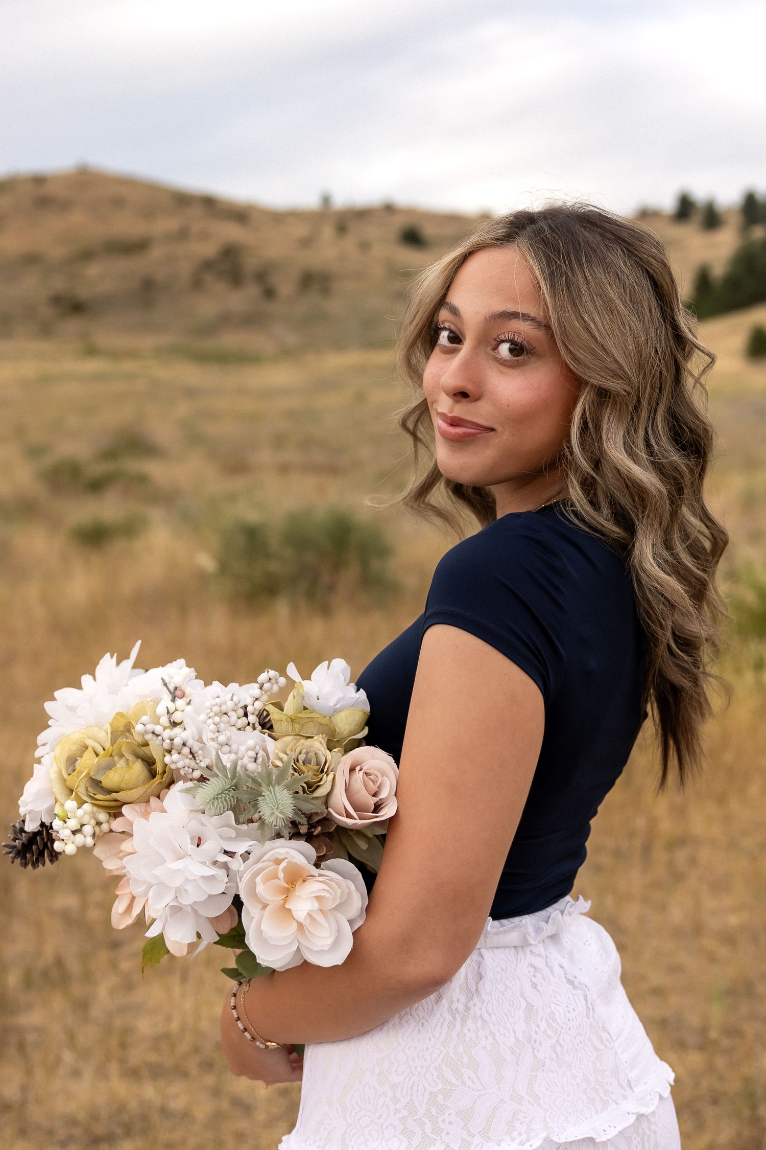 A person in a white lace dress holds a delicate bouquet of white and peach flowers against a golden hillside background.