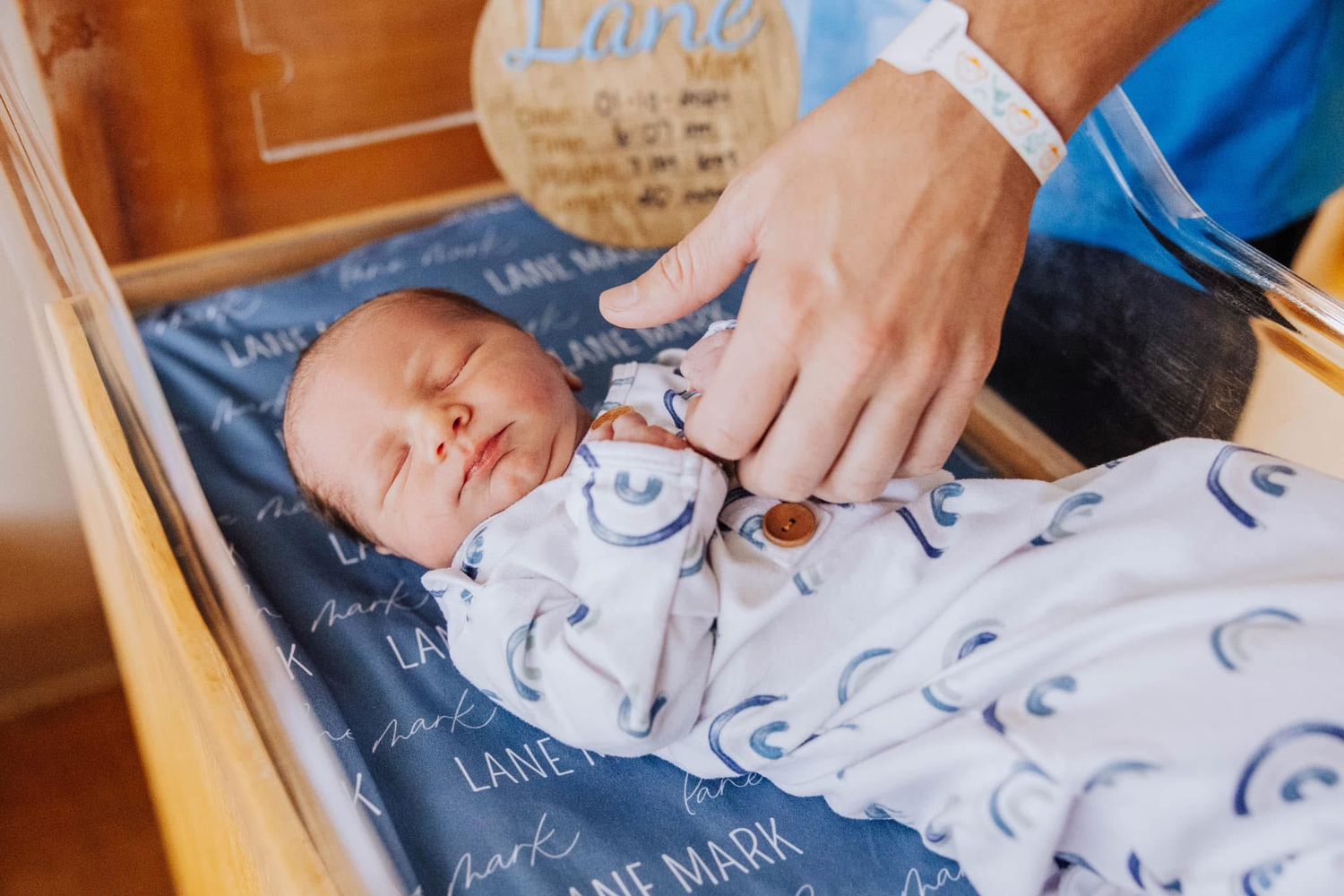 Sleeping newborn baby swaddled in a personalized Lane Mark blanket inside a wooden bassinet.