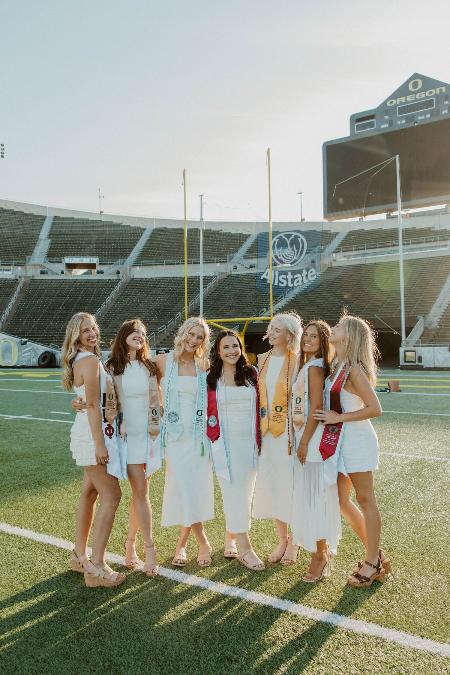 A group in white dresses poses together on a football field at sunset with stadium seating visible in background.