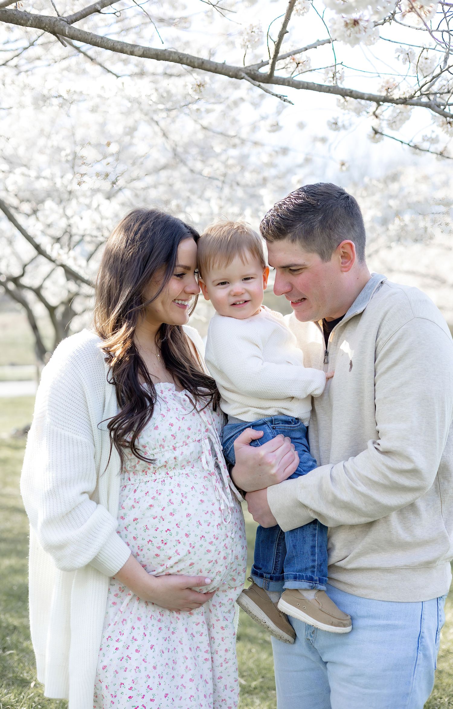 Family of three sharing tender moments under blooming cherry blossom trees during spring photo session.