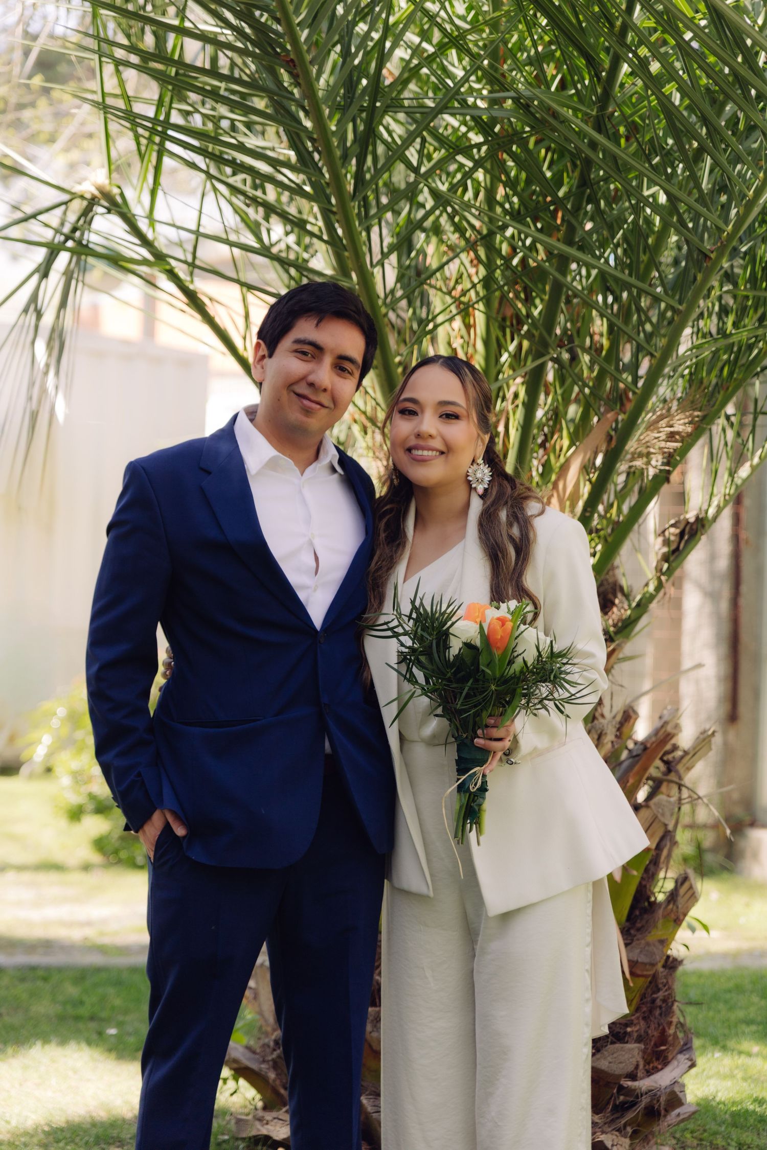 Smiling couple in wedding attire pose before a palm tree, groom in blue suit, bride in white pantsuit holding bouquet.