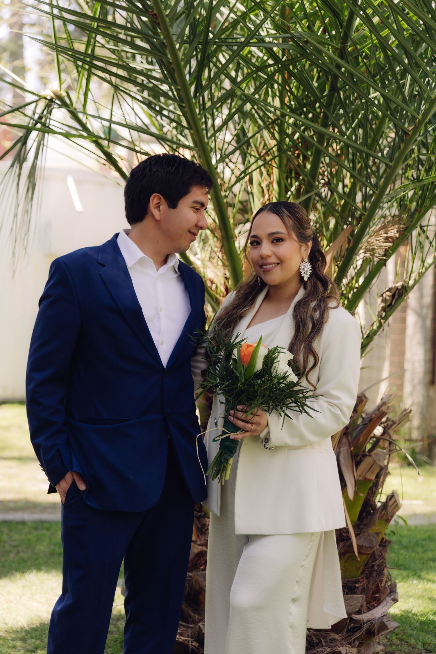 Wedding couple standing by a palm tree, groom in navy suit gazes at bride in white pantsuit holding floral bouquet.