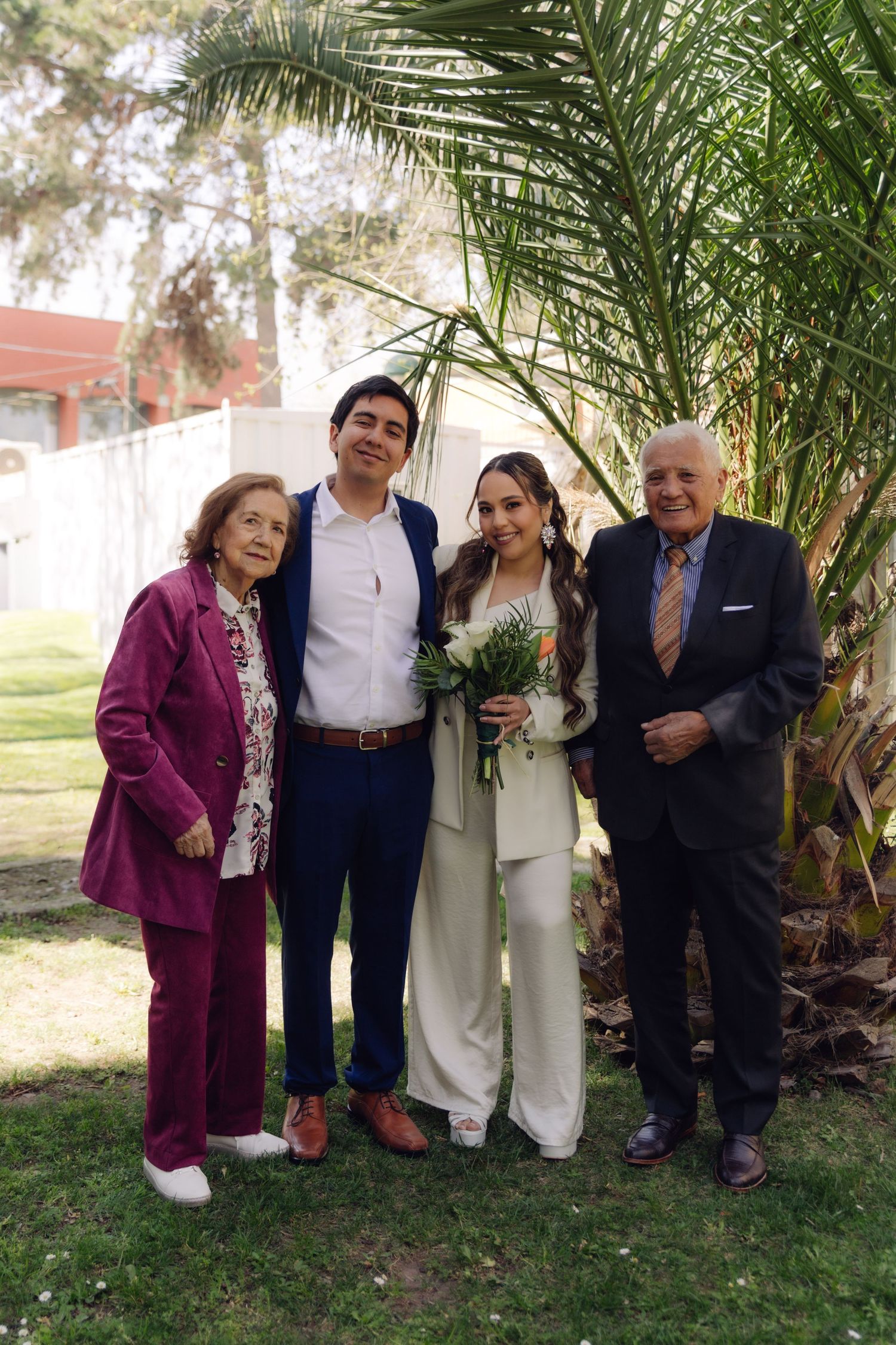 Wedding couple poses with two elderly relatives outdoors near a palm tree, bride holding a green and orange bouquet.