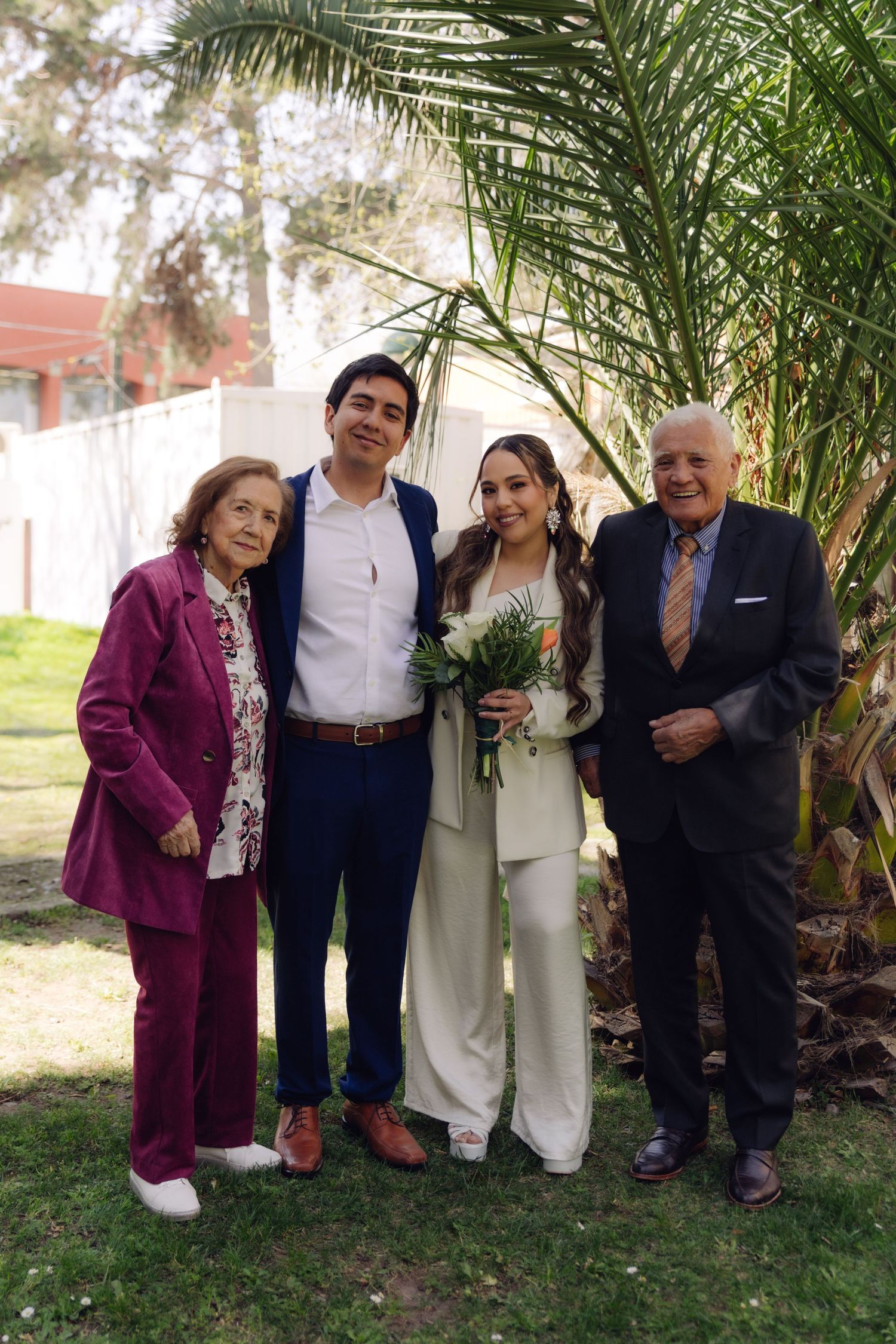 Newlyweds stand with elderly grandparents near a palm tree, bride in white pantsuit holding a colorful bouquet.