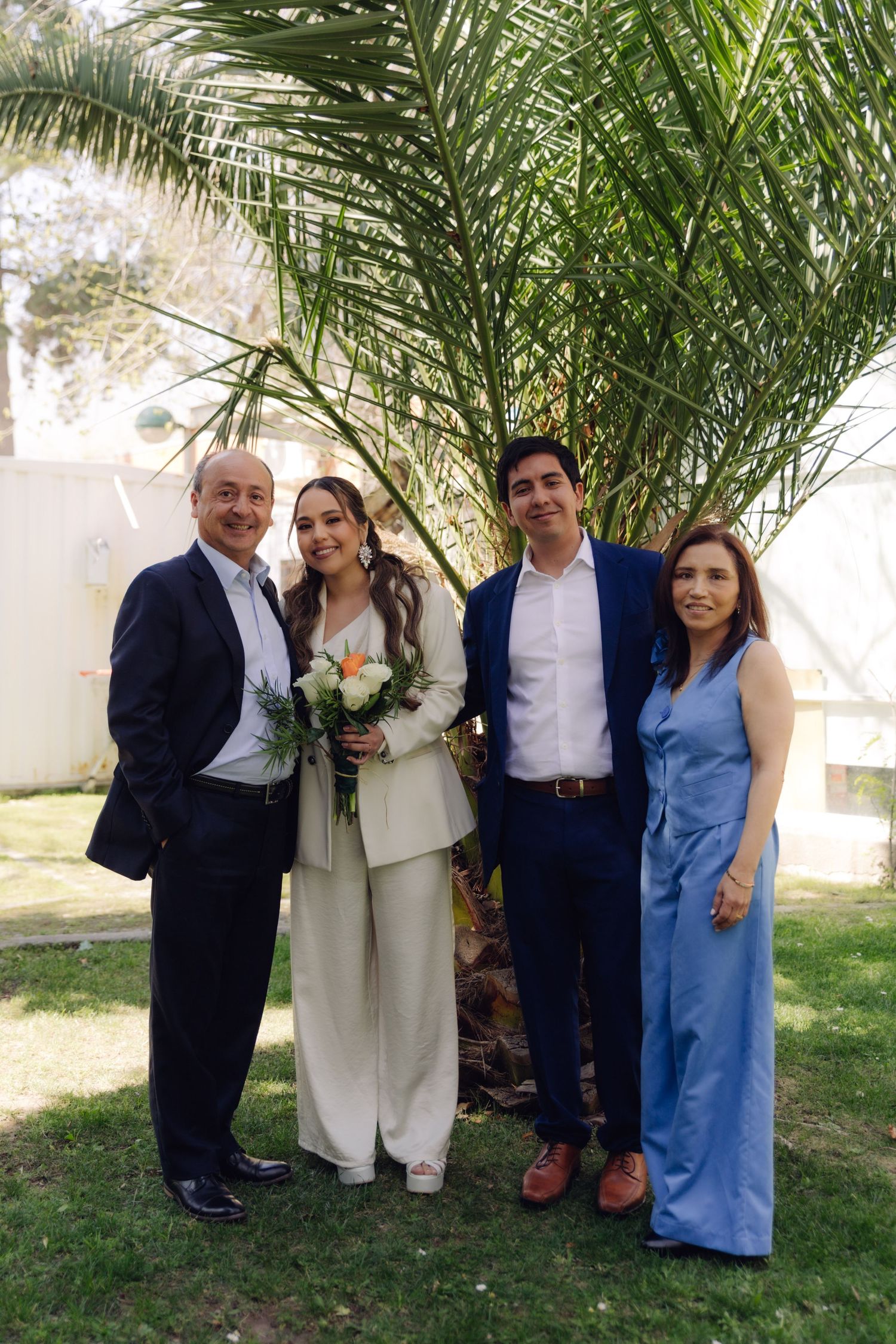 Wedding couple poses with parents outdoors, bride in white pantsuit with bouquet, father in black suit, mother in blue.