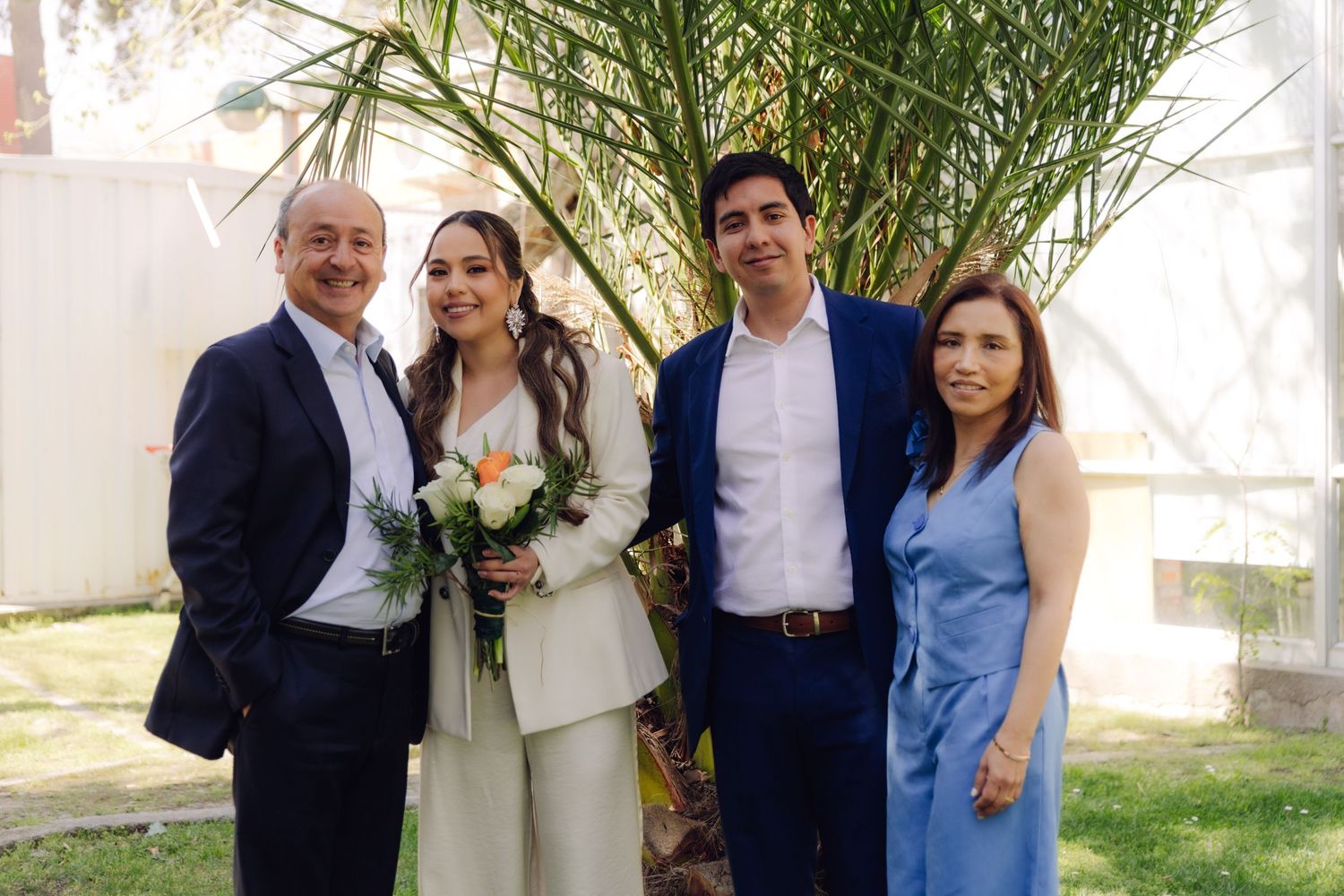 Bride in white pantsuit with bouquet stands between parents and groom in blue suit near a palm tree outdoors.