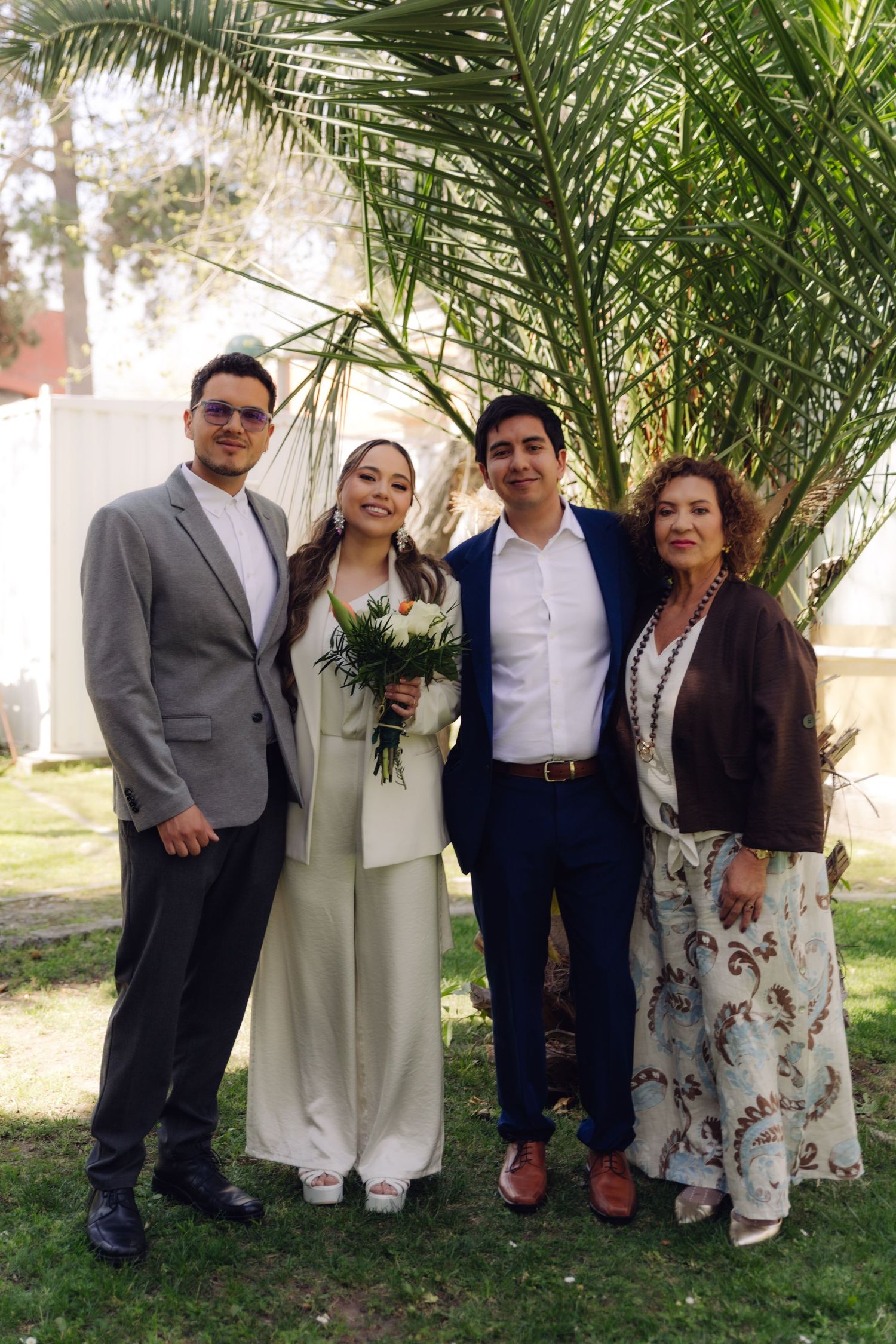Wedding couple with two family members pose outdoors near a palm tree, bride holding bouquet in white pantsuit.
