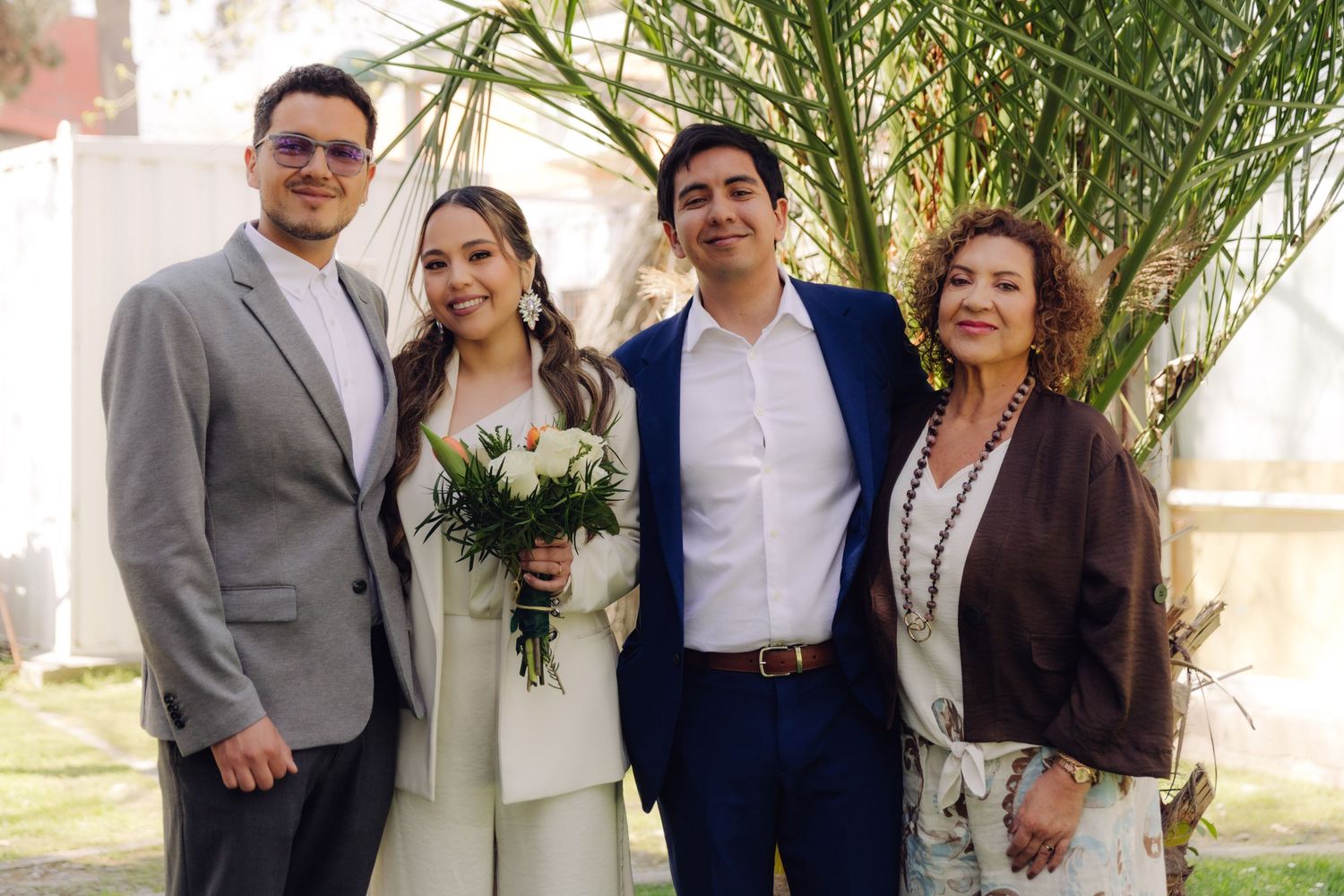 Smiling wedding group of four poses near a palm tree, bride in white pantsuit holding a white and orange bouquet.