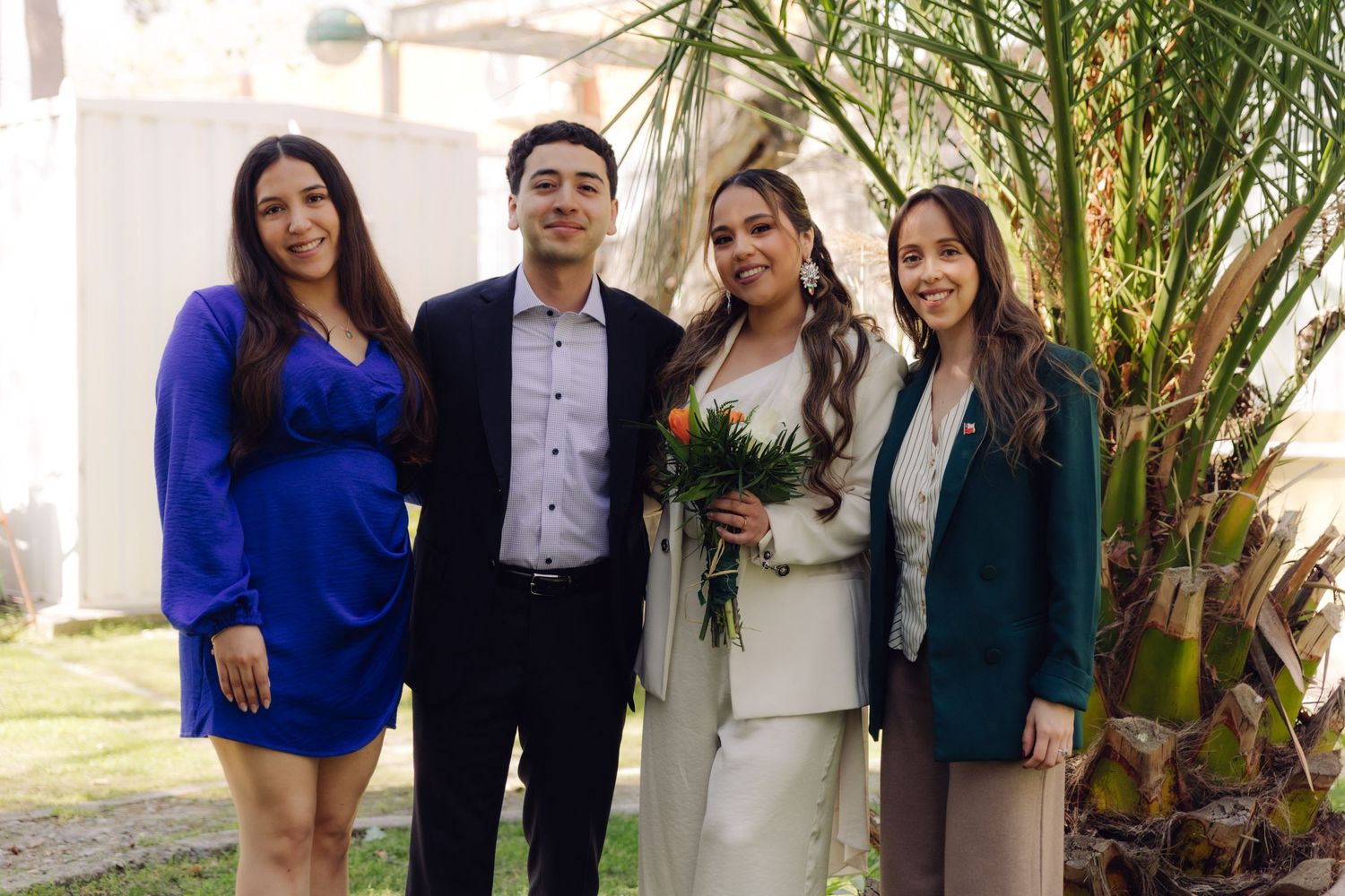 Bride in white pantsuit with orange bouquet poses with groom and two women in blue and teal outfits near a palm tree.