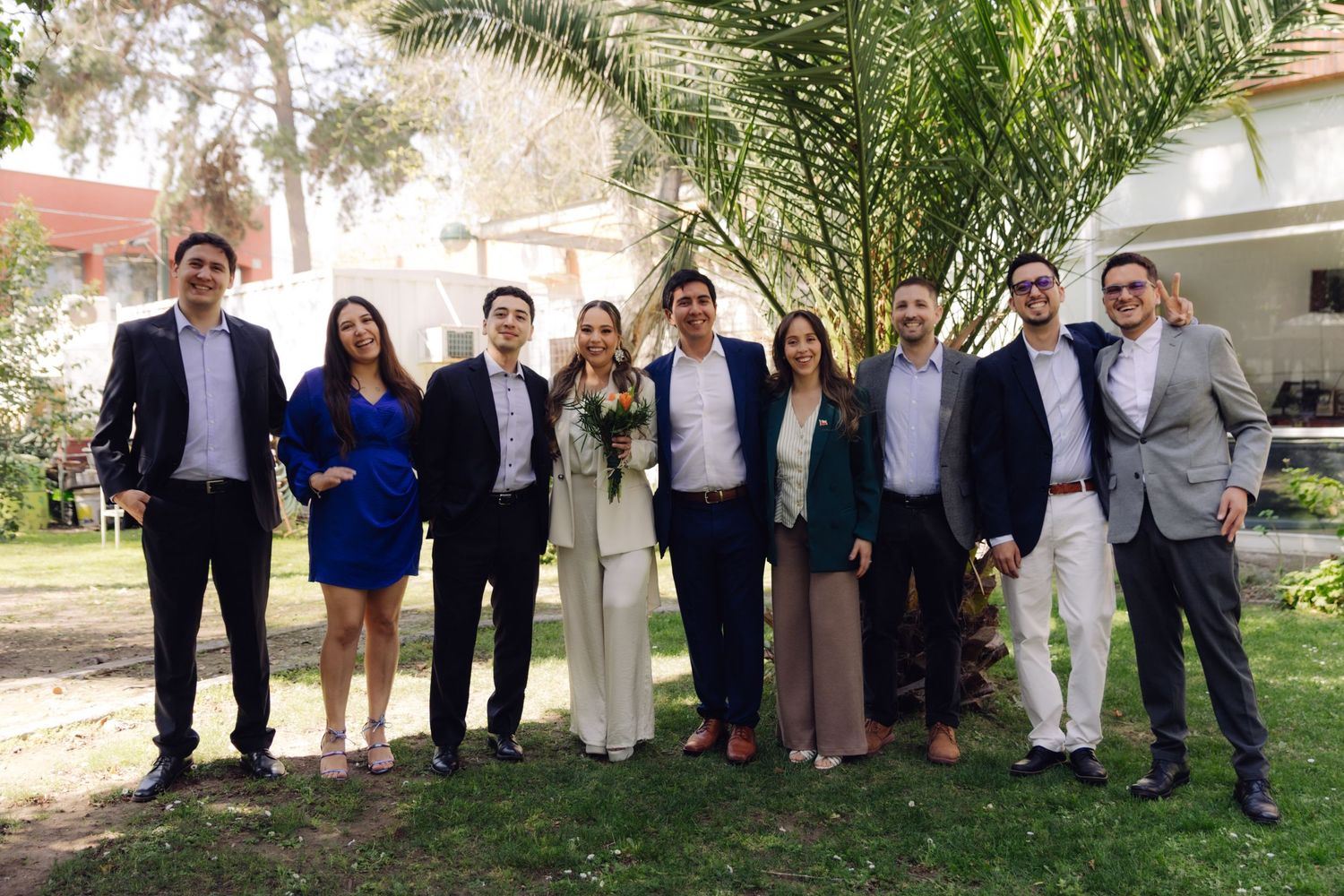 Group of elegantly dressed wedding guests posing together outdoors in a garden setting.