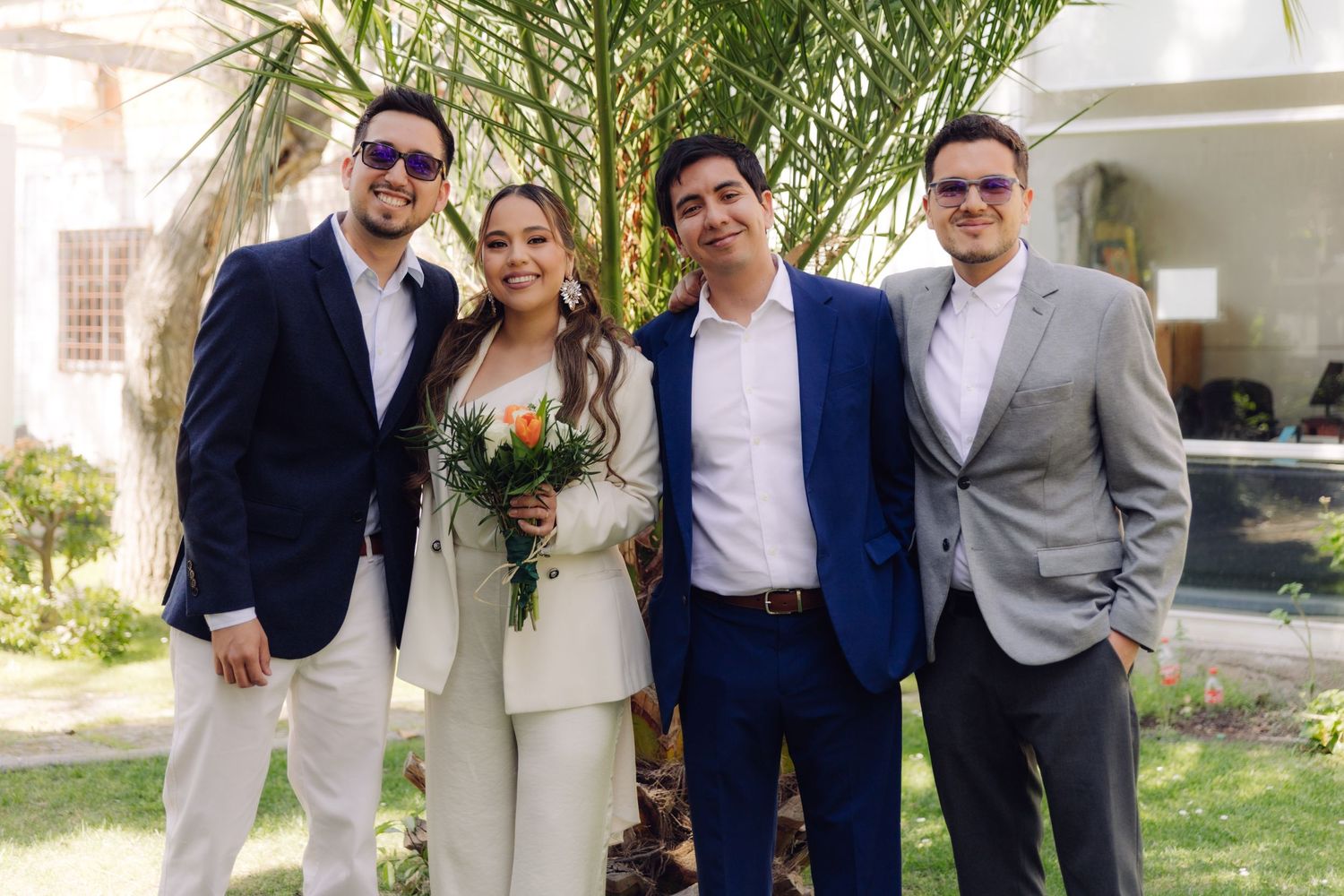 Bride in white suit holding bouquet poses with three formally dressed men outdoors.