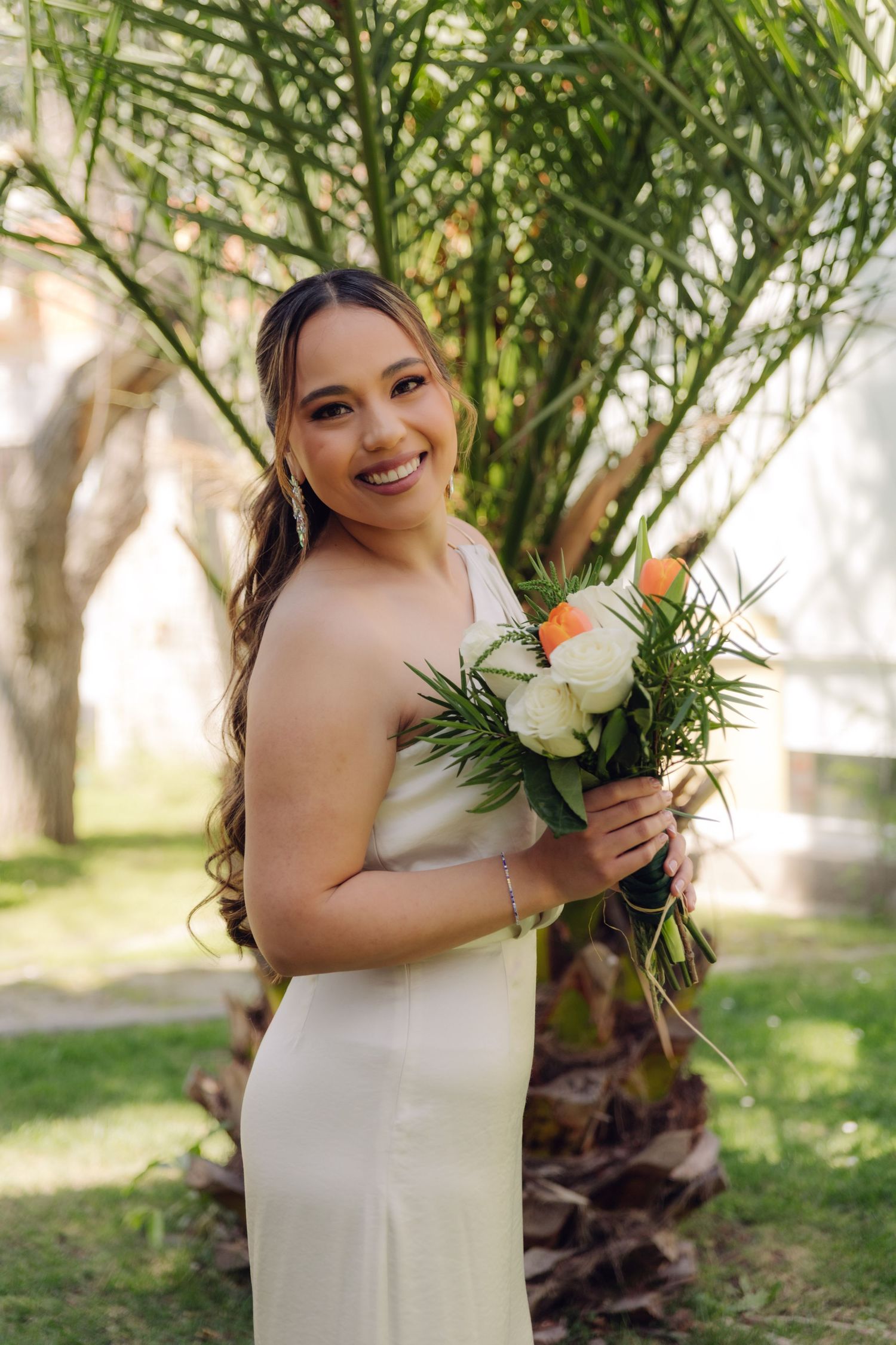 Radiant bride in white dress holds orange and white bouquet beside a tropical palm tree.