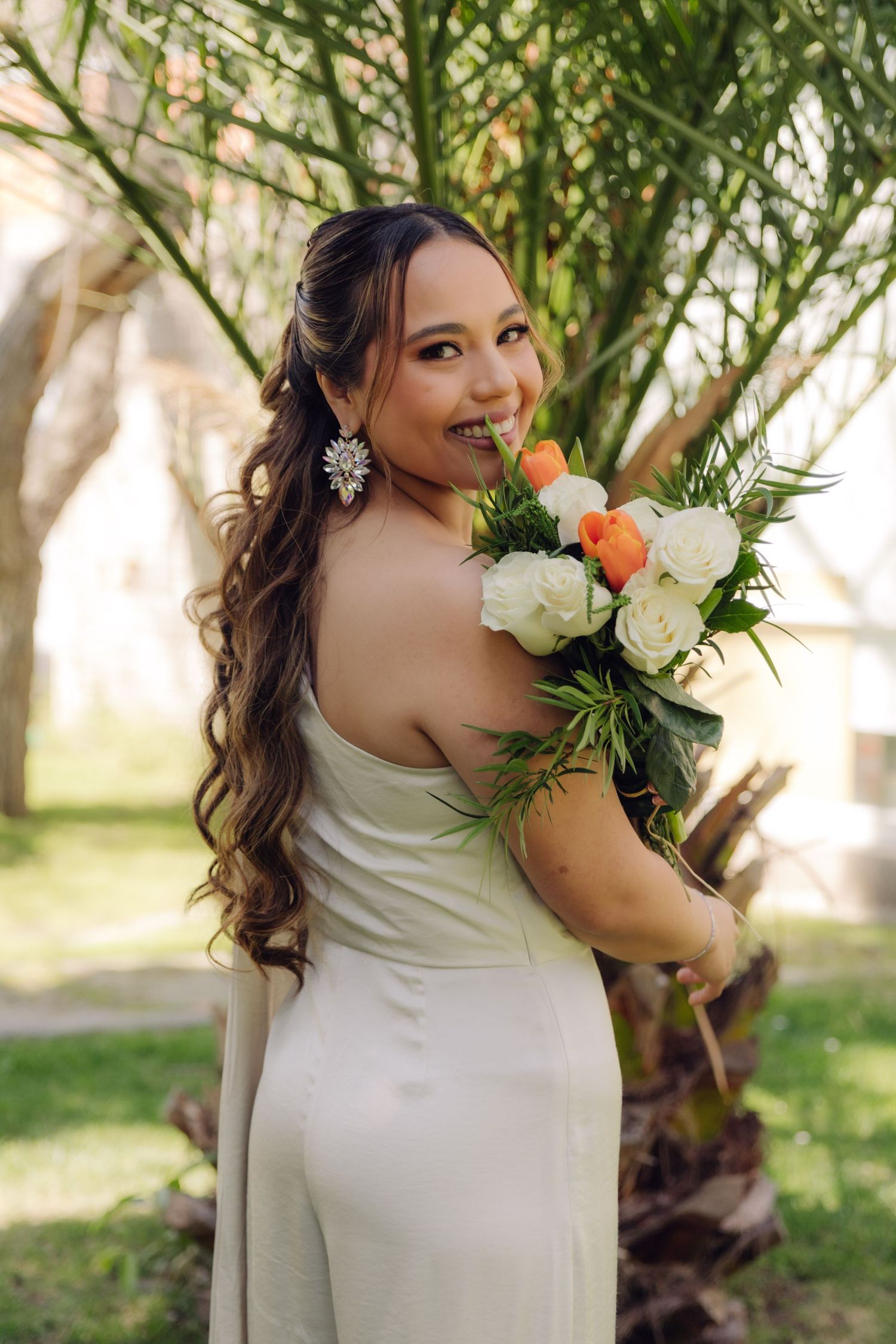 Elegant bride in white gown smiles over shoulder holding bouquet near a palm tree.