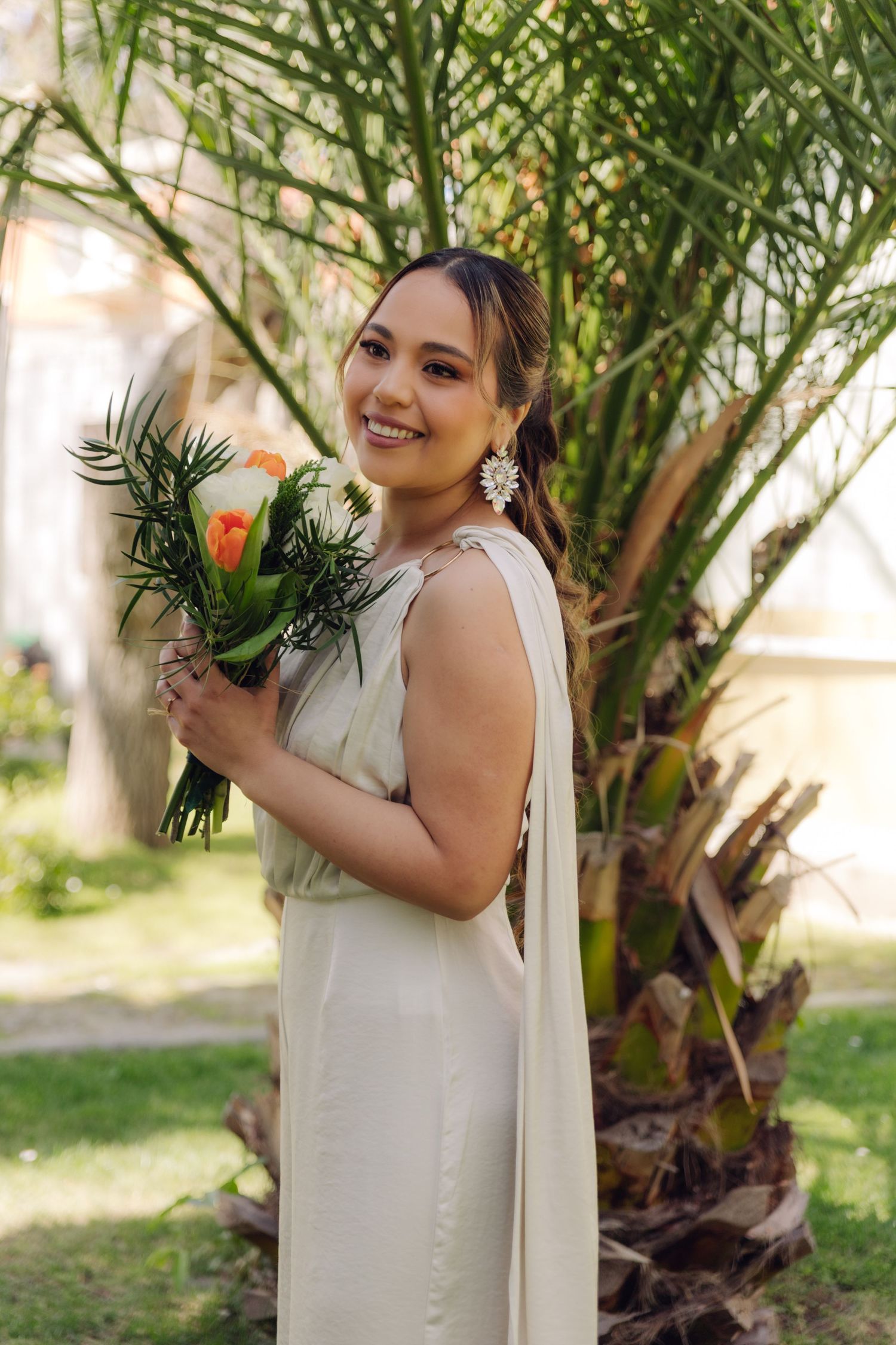 Smiling bride in white dress holds colorful bouquet while standing beside a lush palm tree.