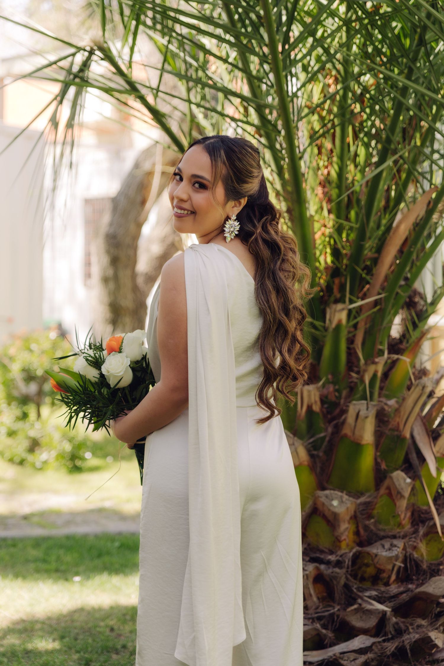 Beautiful bride in white jumpsuit with cape holds bouquet while posing near tropical palms.