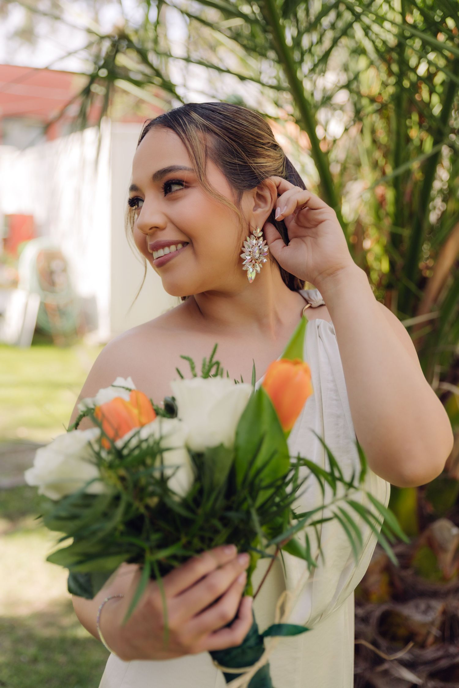 Glowing bride adjusts sparkling earring while holding orange and white bouquet outdoors.