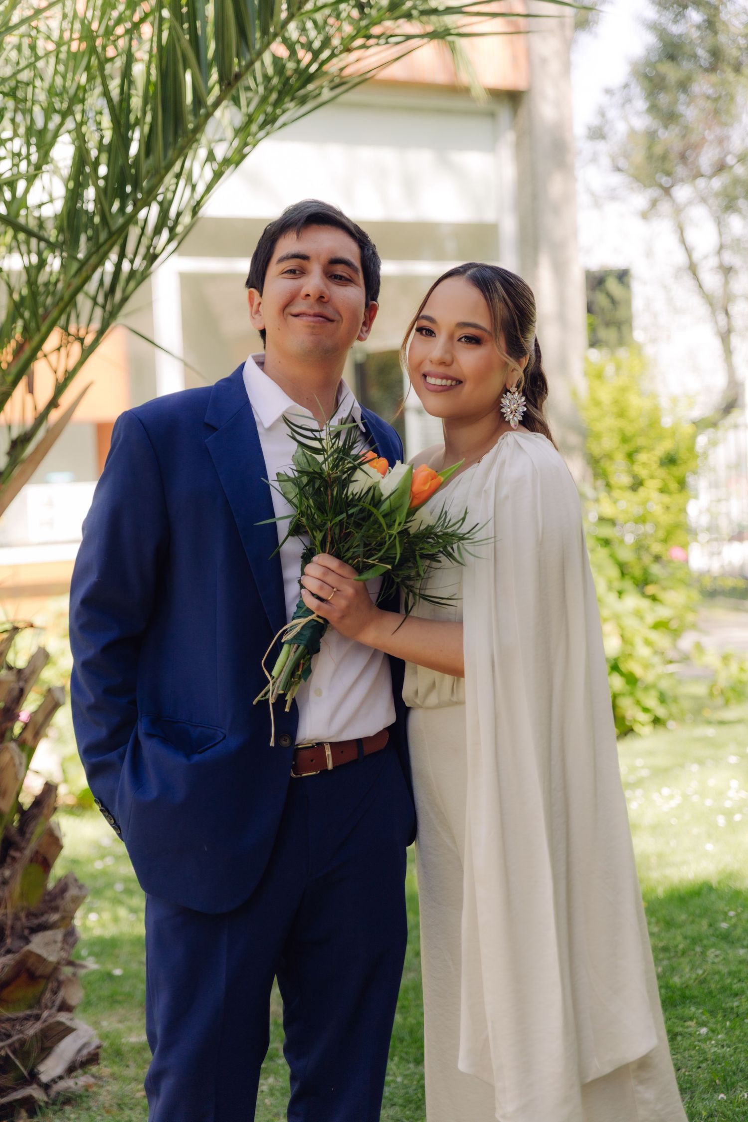 Smiling bride and groom in blue suit pose together holding bouquet in garden setting.