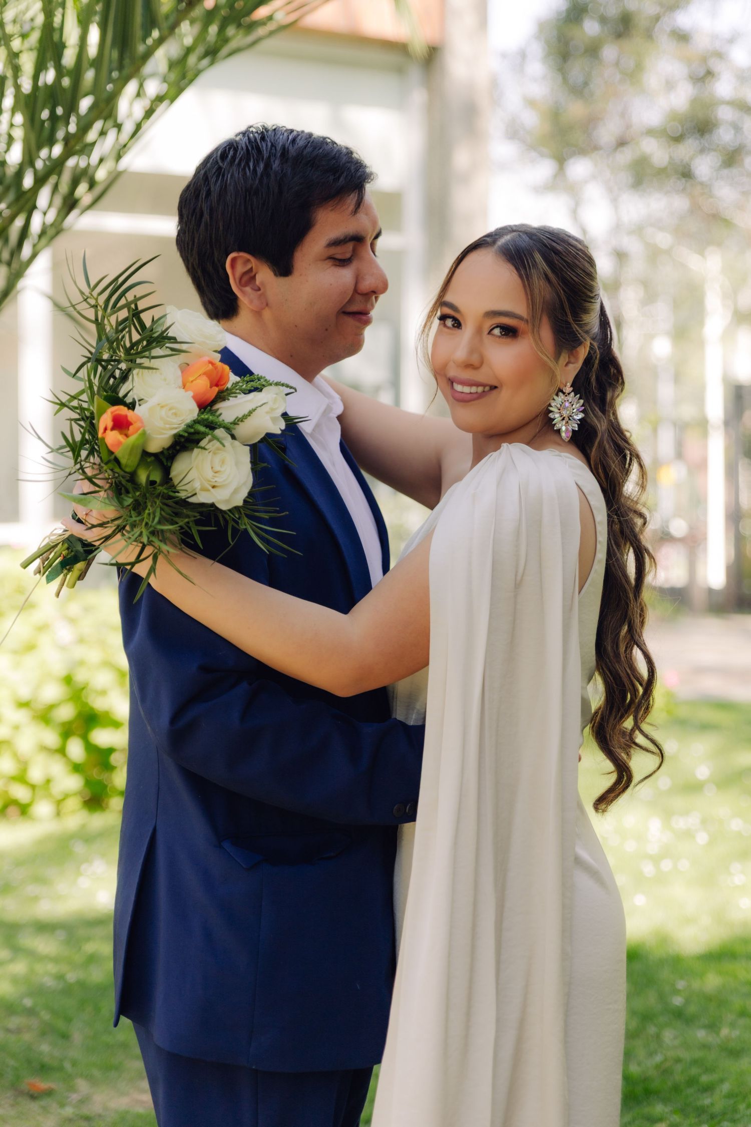 Groom in blue suit embraces smiling bride holding floral bouquet in a sunlit garden.