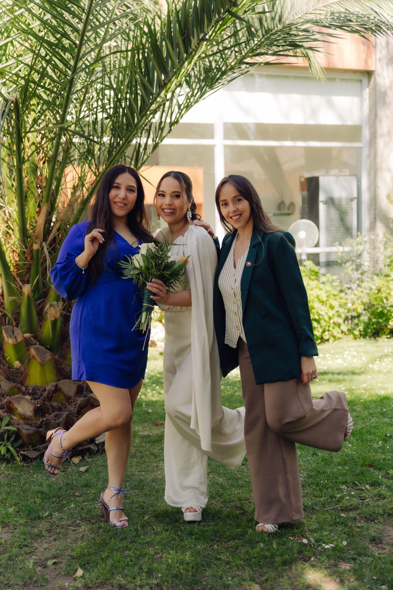 Three women pose outdoors at a wedding, bride in white holding bouquet flanked by friends in blue dress and green blazer.