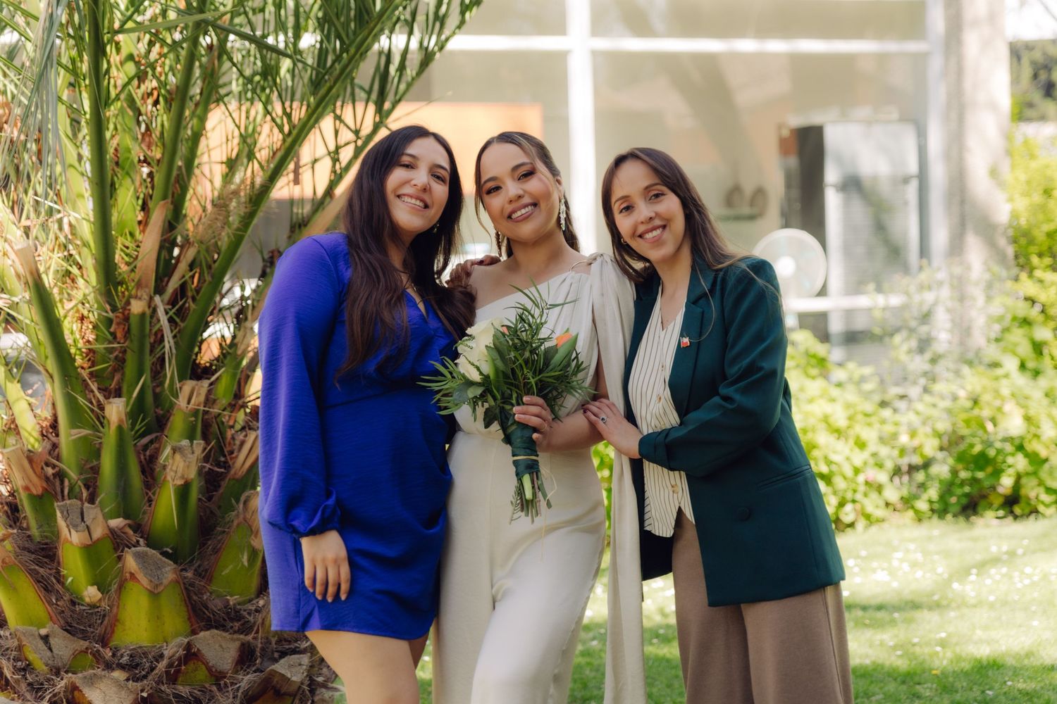Bride in white jumpsuit holds bouquet while posing with two friends in blue dress and green blazer at outdoor wedding.