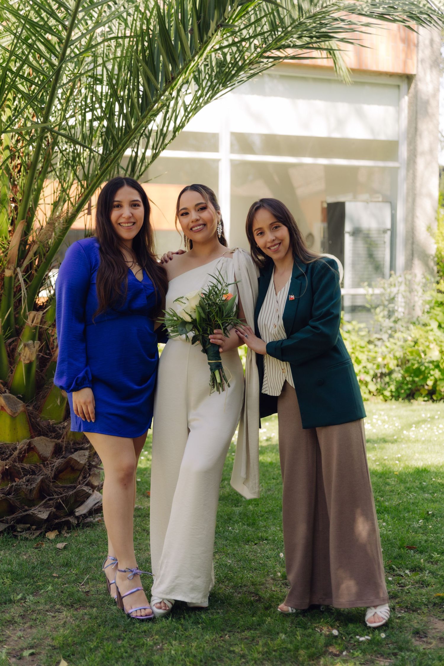 Three women smile together at a wedding outdoors, bride in white holding flowers between guests in blue and green outfits.