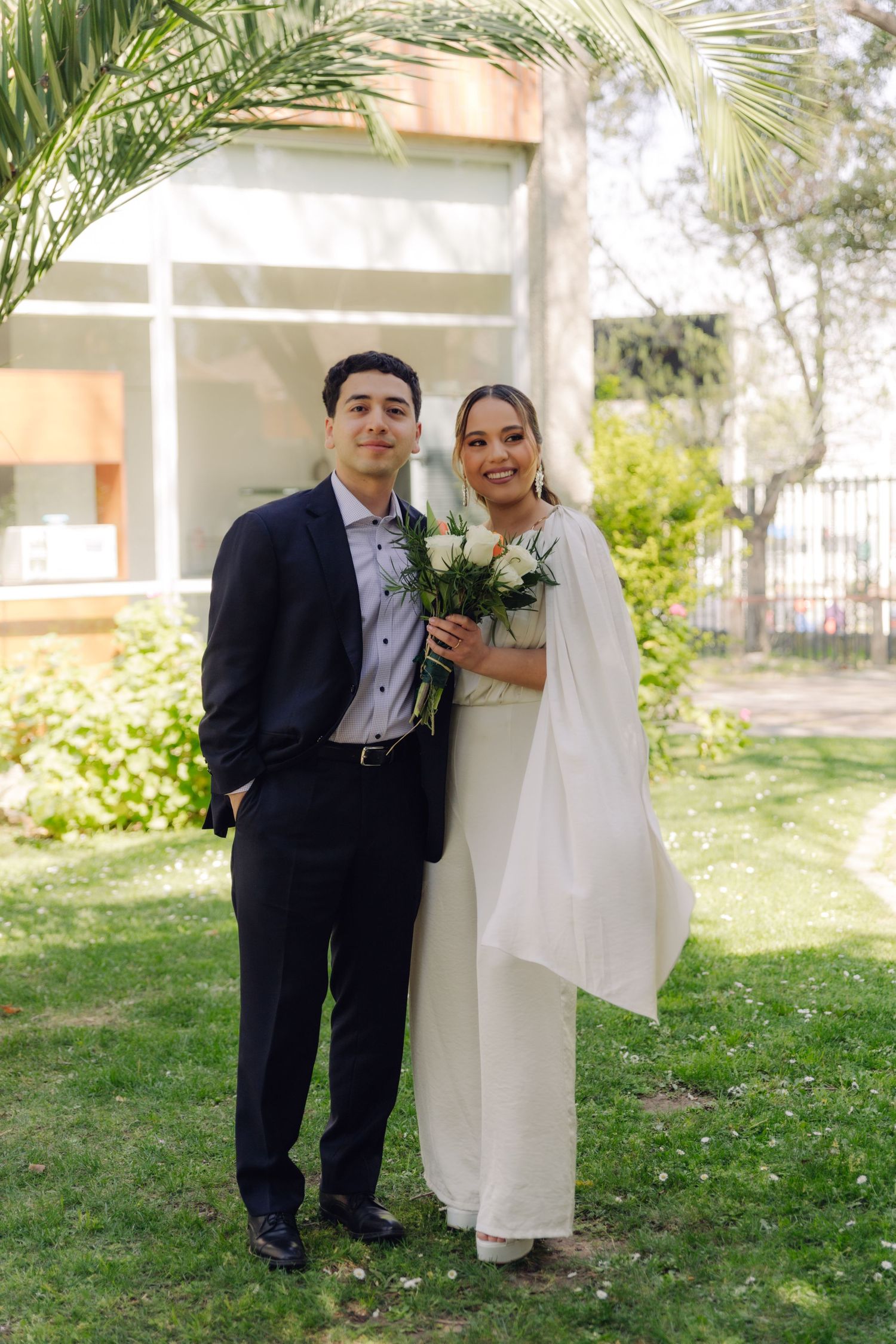 Newlywed couple poses in garden, groom in navy suit beside bride in white dress holding rose bouquet at civil wedding.