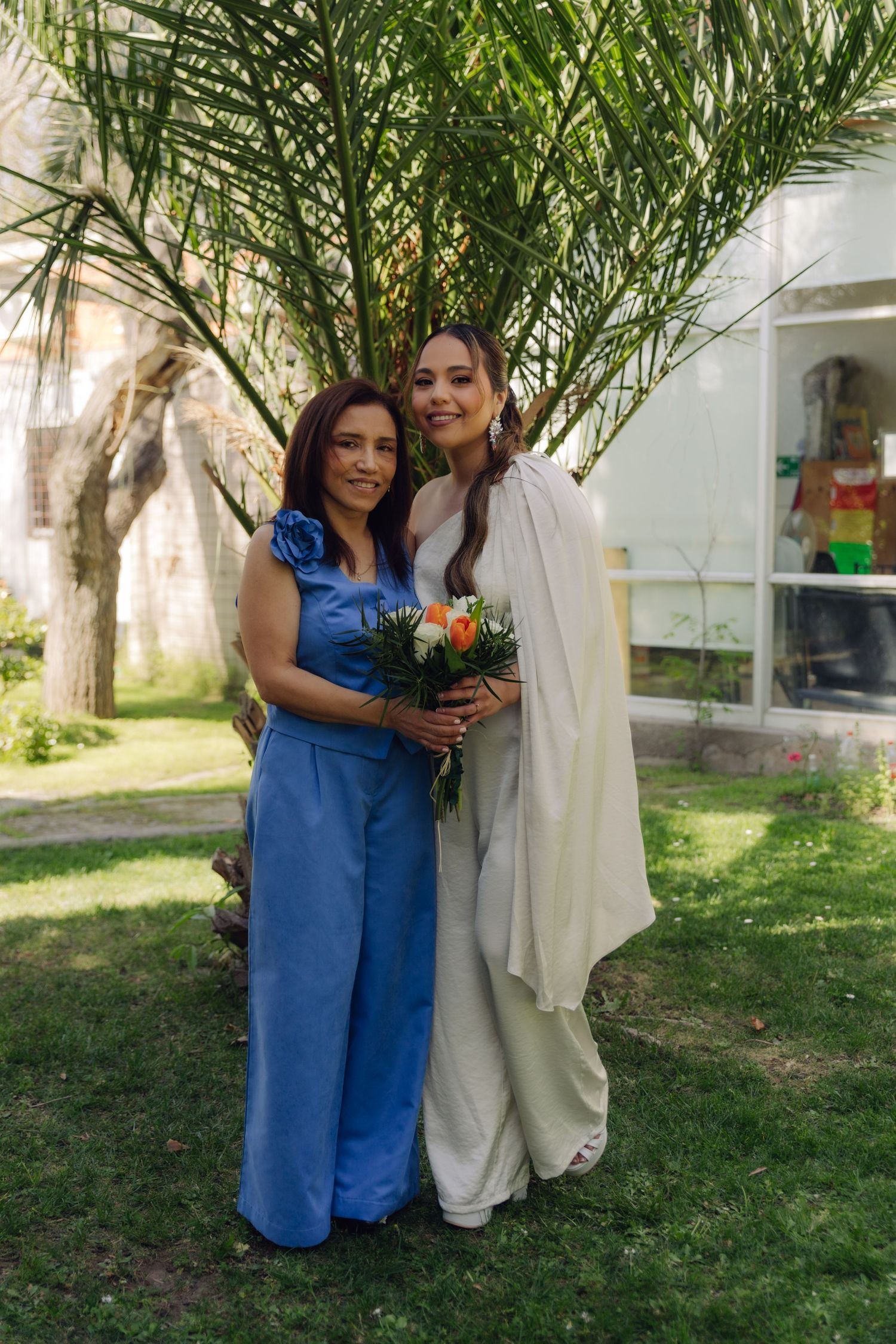 Bride in white dress with cape holds colorful bouquet while posing with older woman in blue jumpsuit at wedding.