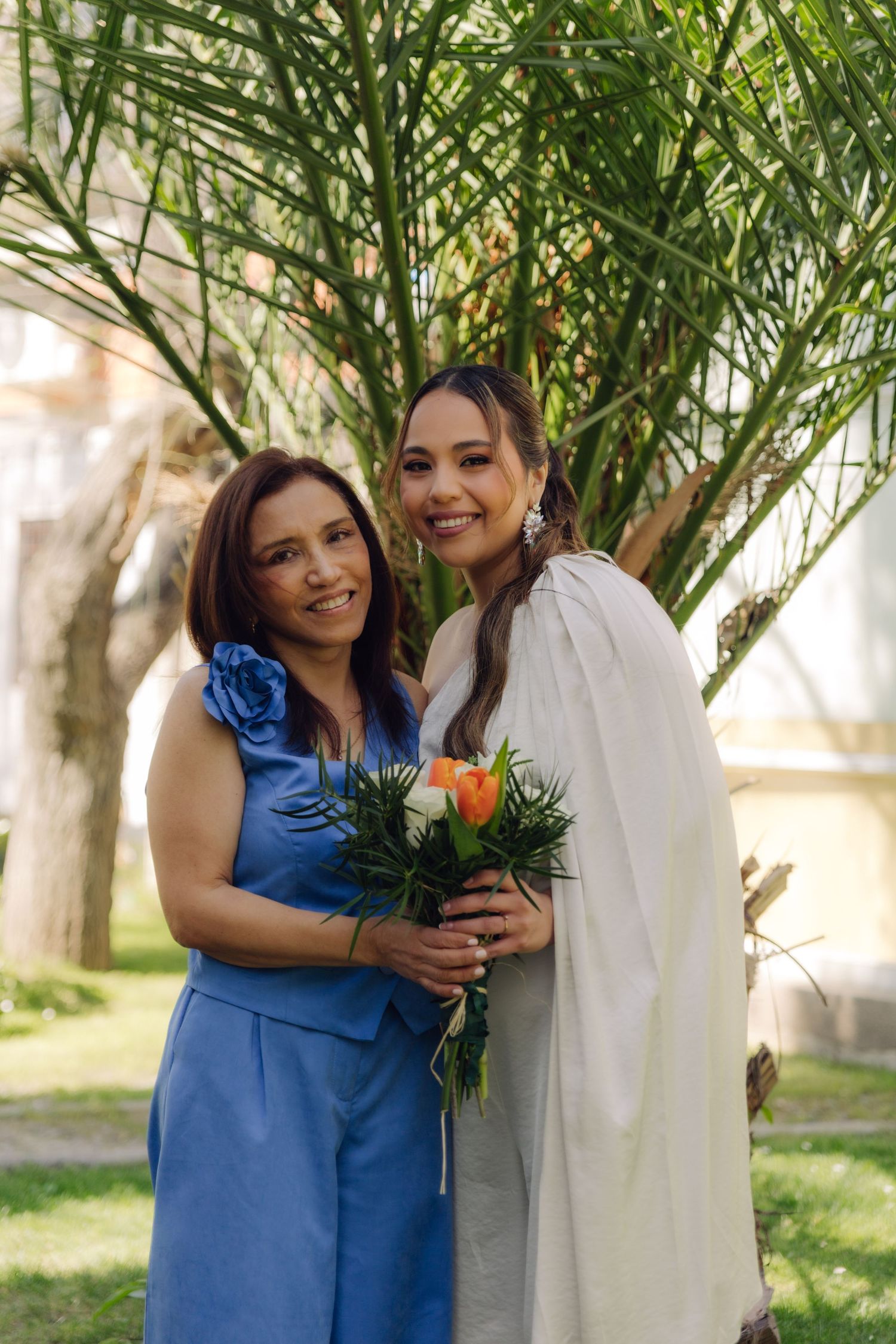 Smiling bride in white dress and cape holds orange and white bouquet with older woman in blue dress at outdoor wedding.