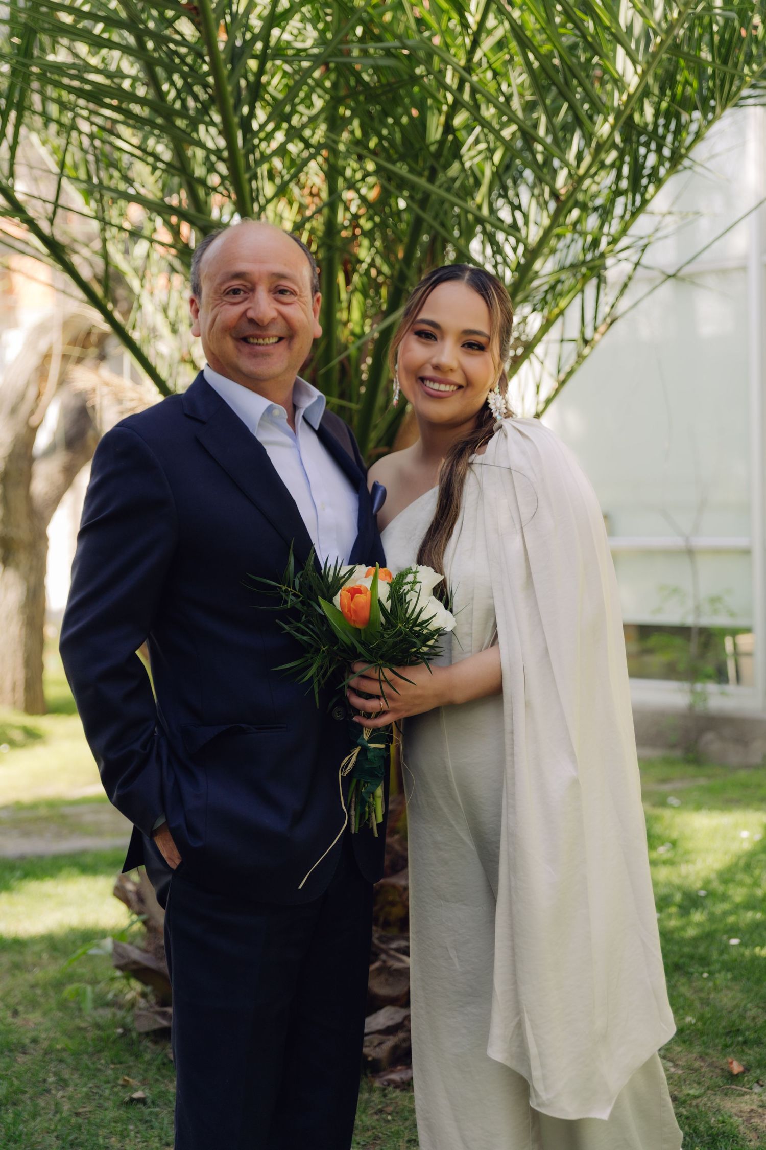 Older man in navy suit smiles beside bride in white dress holding colorful bouquet at outdoor wedding ceremony.