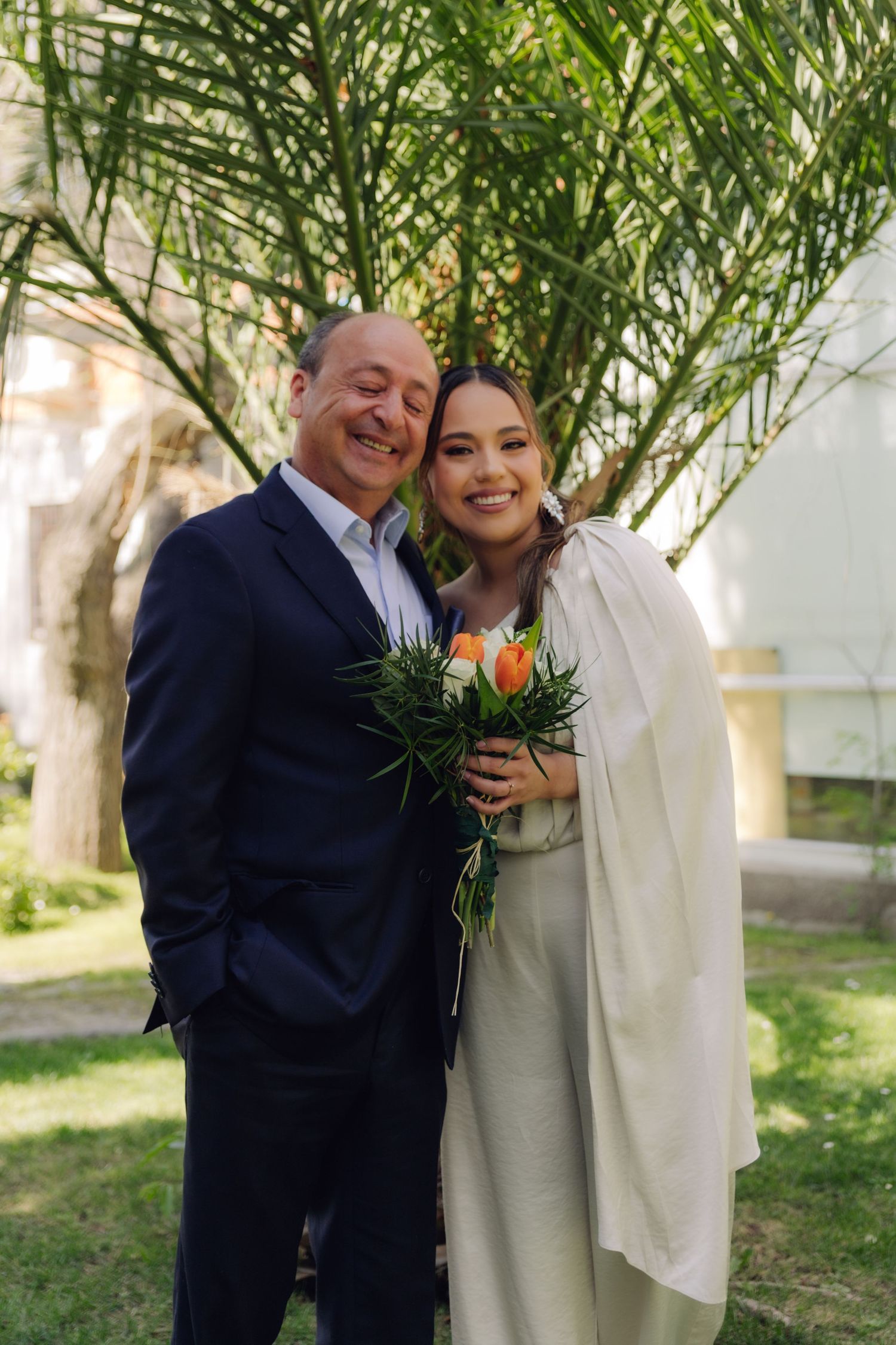 Bride in white dress leans on older man in navy suit while holding orange and green bouquet at outdoor wedding.
