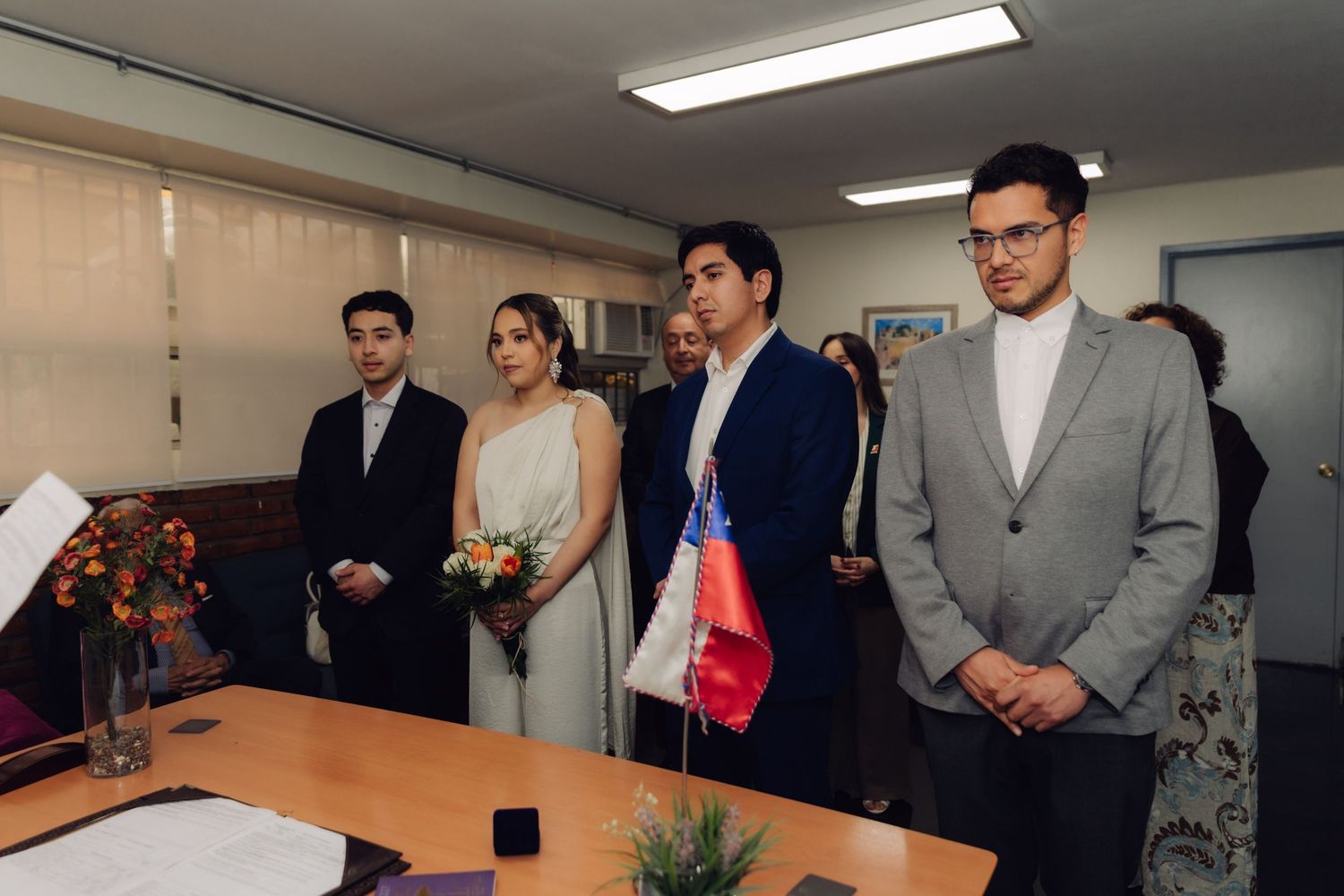 Wedding party stands at civil ceremony indoors with Chilean flag, bride in white holding flowers with groom and witnesses.