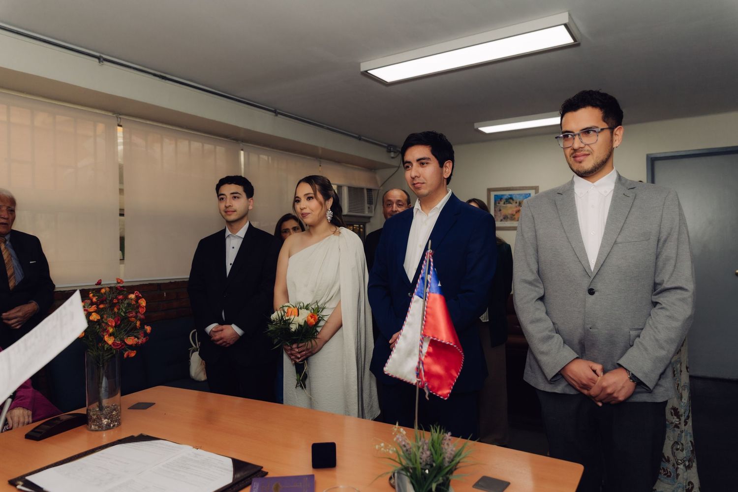 Bride and groom stand with witnesses at indoor civil wedding ceremony beside Chilean flag in an office setting.