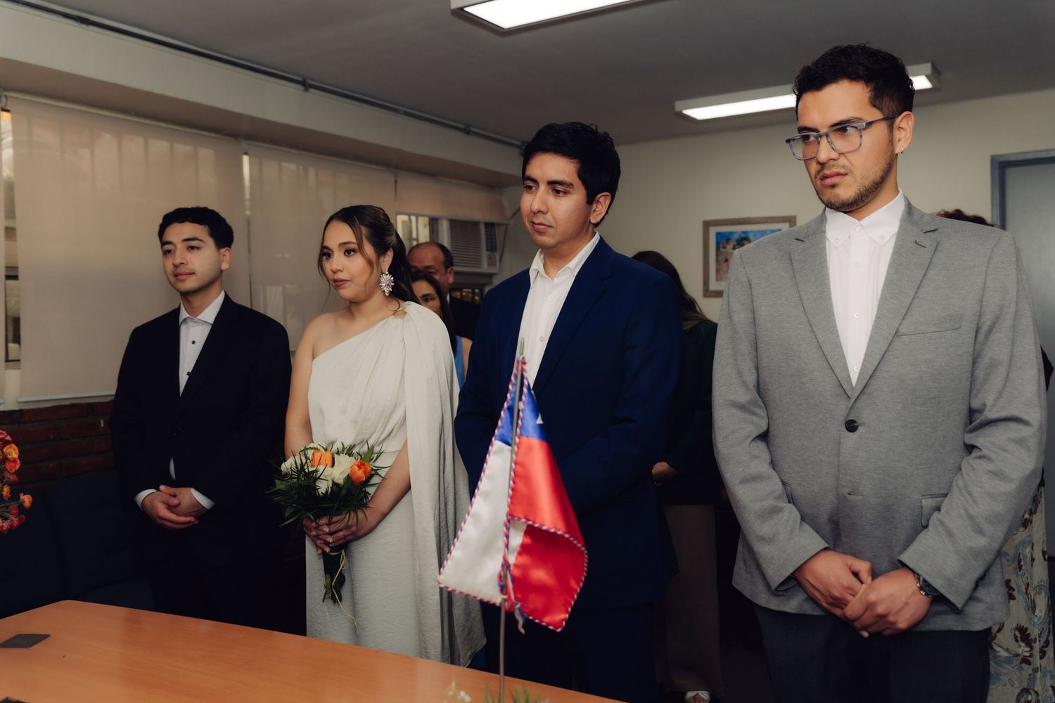 Couple stands at civil wedding ceremony with Chilean flag and witnesses in formal attire.