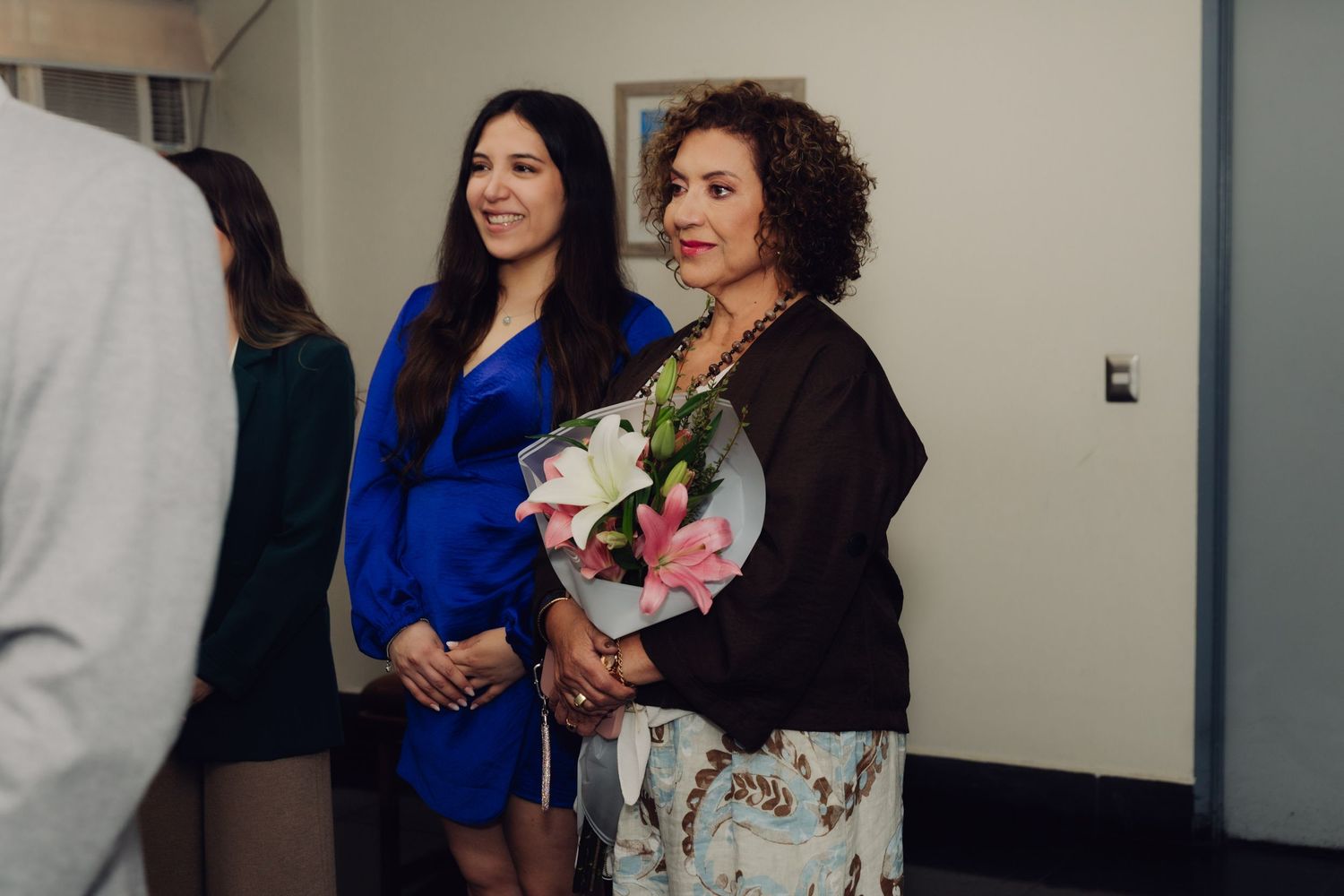 Two women smile holding flowers at a wedding celebration, one in blue dress.