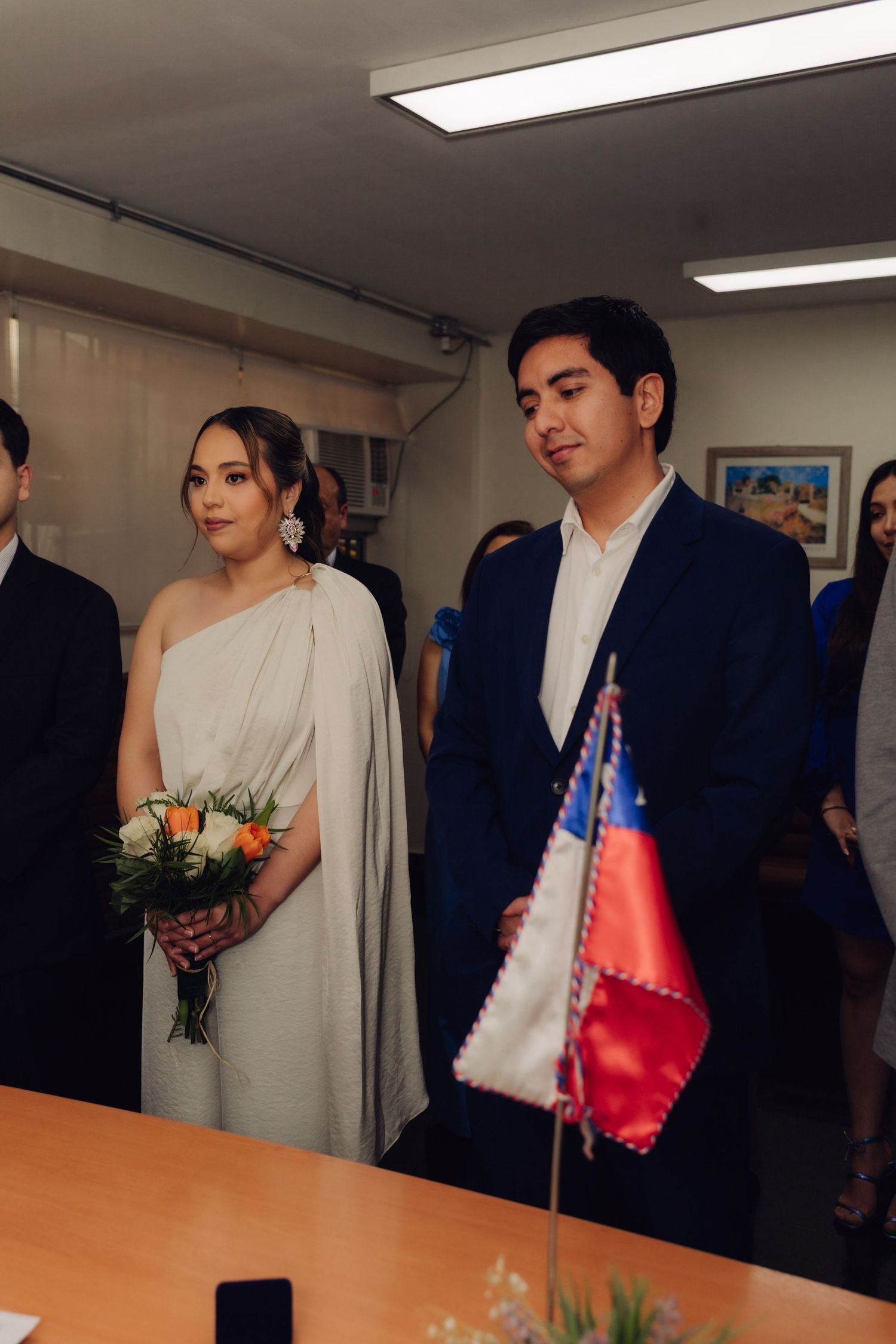 Bride and groom stand solemnly before Chilean flag during civil wedding ceremony.